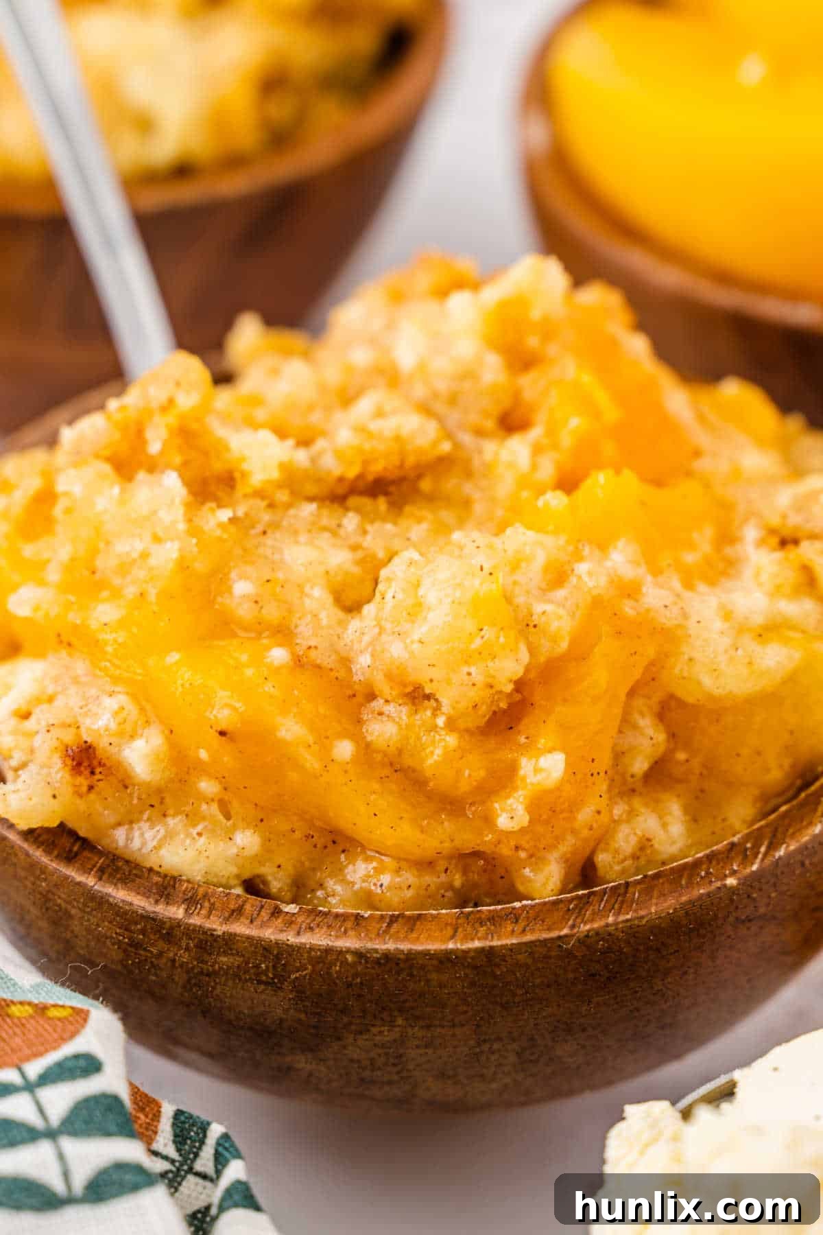 Effortless Peach Cobbler Cake 2 A serving of golden-brown peach cobbler dump cake in a white bowl, topped with a scoop of melting vanilla ice cream and a spoon resting in the dish.