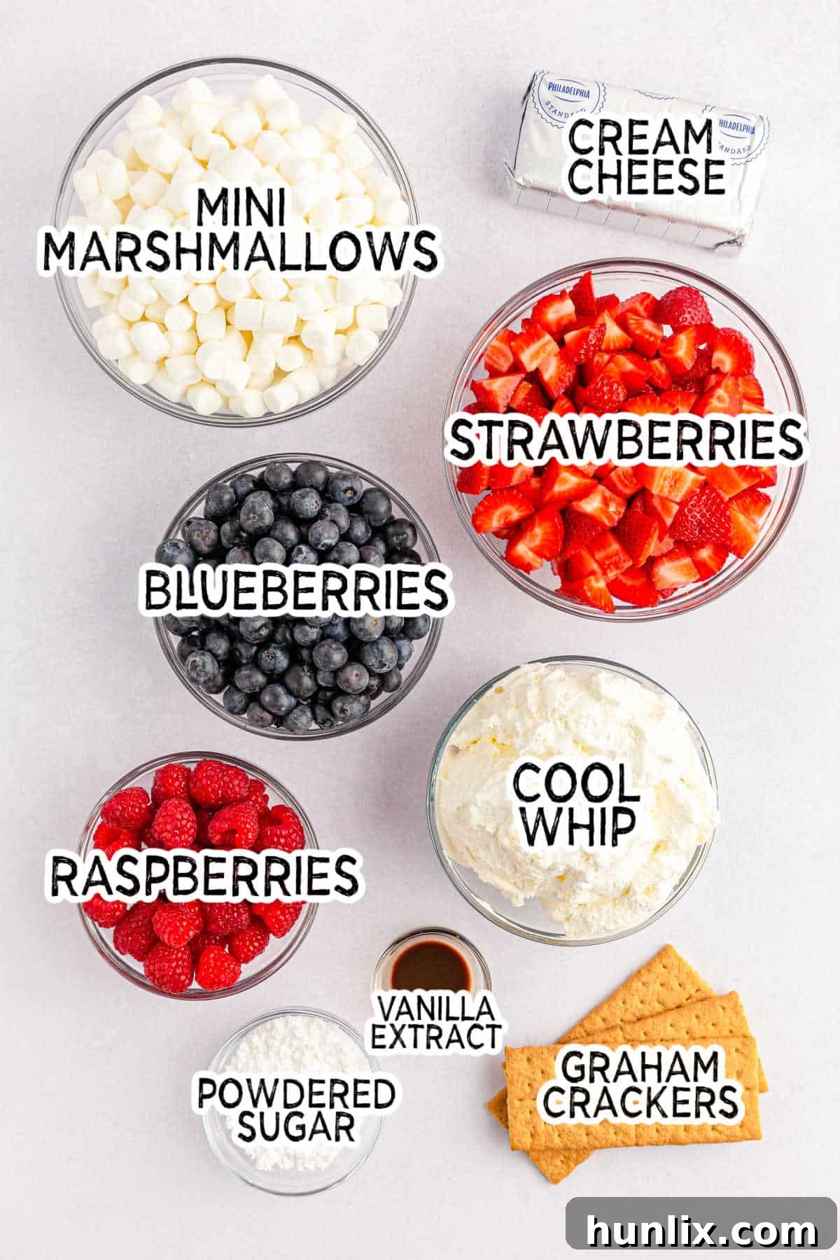 A collection of ingredients laid out, including blocks of cream cheese, a bag of powdered sugar, vanilla extract, a tub of Cool Whip, a bag of mini marshmallows, and bowls of fresh blueberries, diced strawberries, and raspberries.