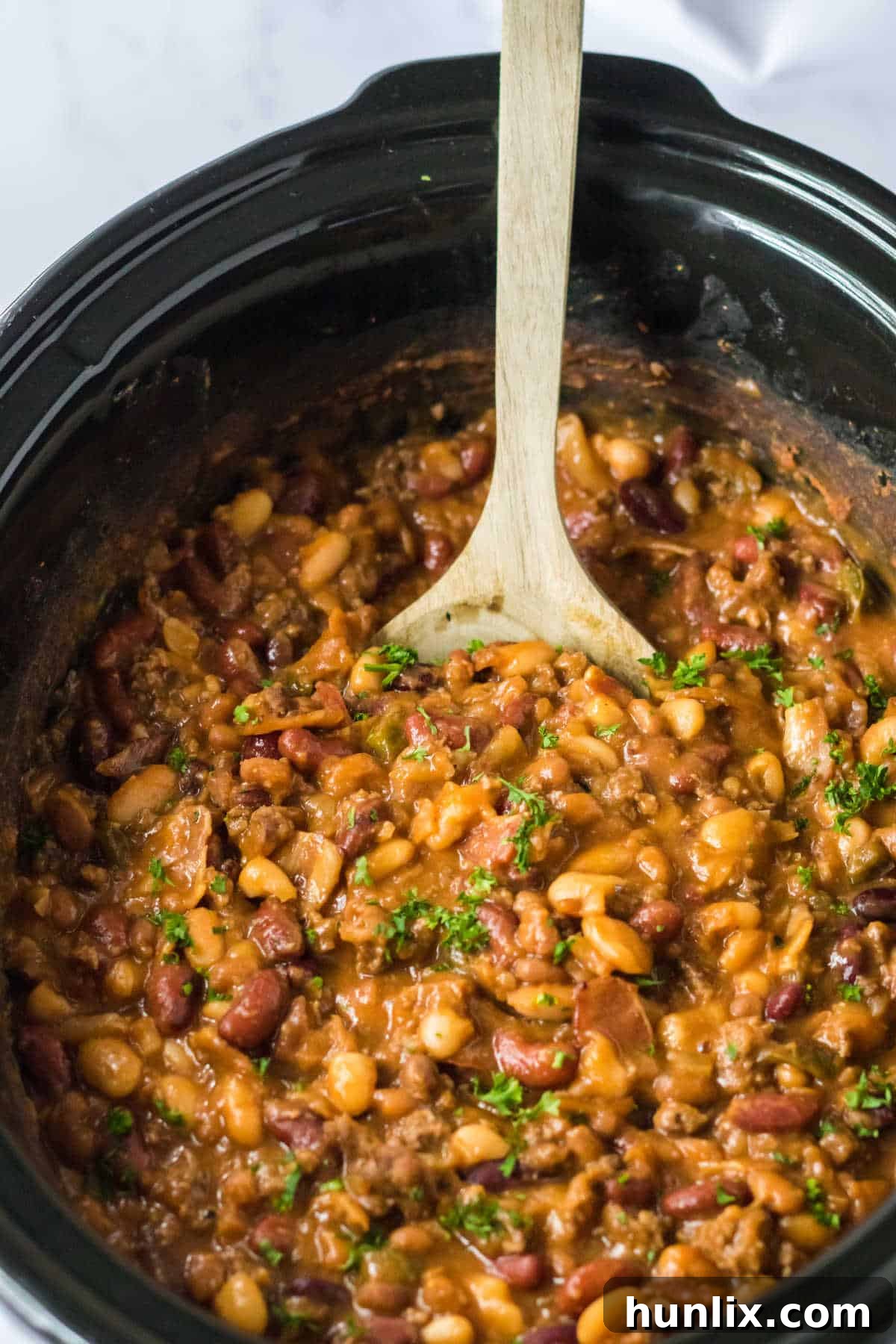 A slow cooker filled with hearty cowboy beans, with a wooden spoon resting in it, ready to serve.