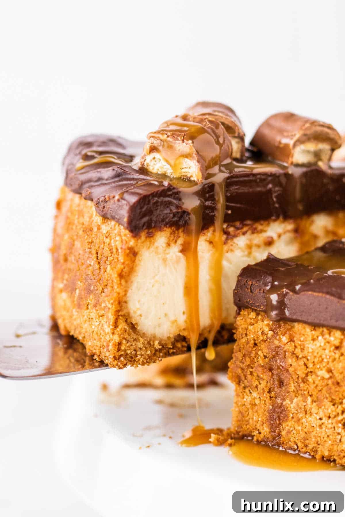 Twix Caramel Crunch Cheesecake 8 A spatula lifting a perfect slice of Twix Cheesecake from the pan, revealing its creamy layers, graham cracker crust, and rich chocolate ganache topping, ready to be served.
