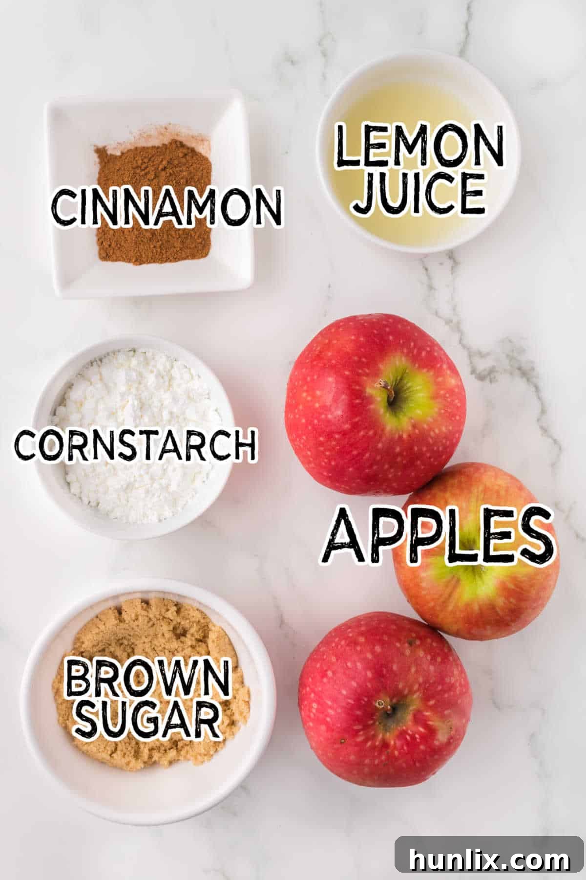 Ingredients for air fryer apples laid out on a white marble counter, including apples, cornstarch, lemon juice, brown sugar, and cinnamon.