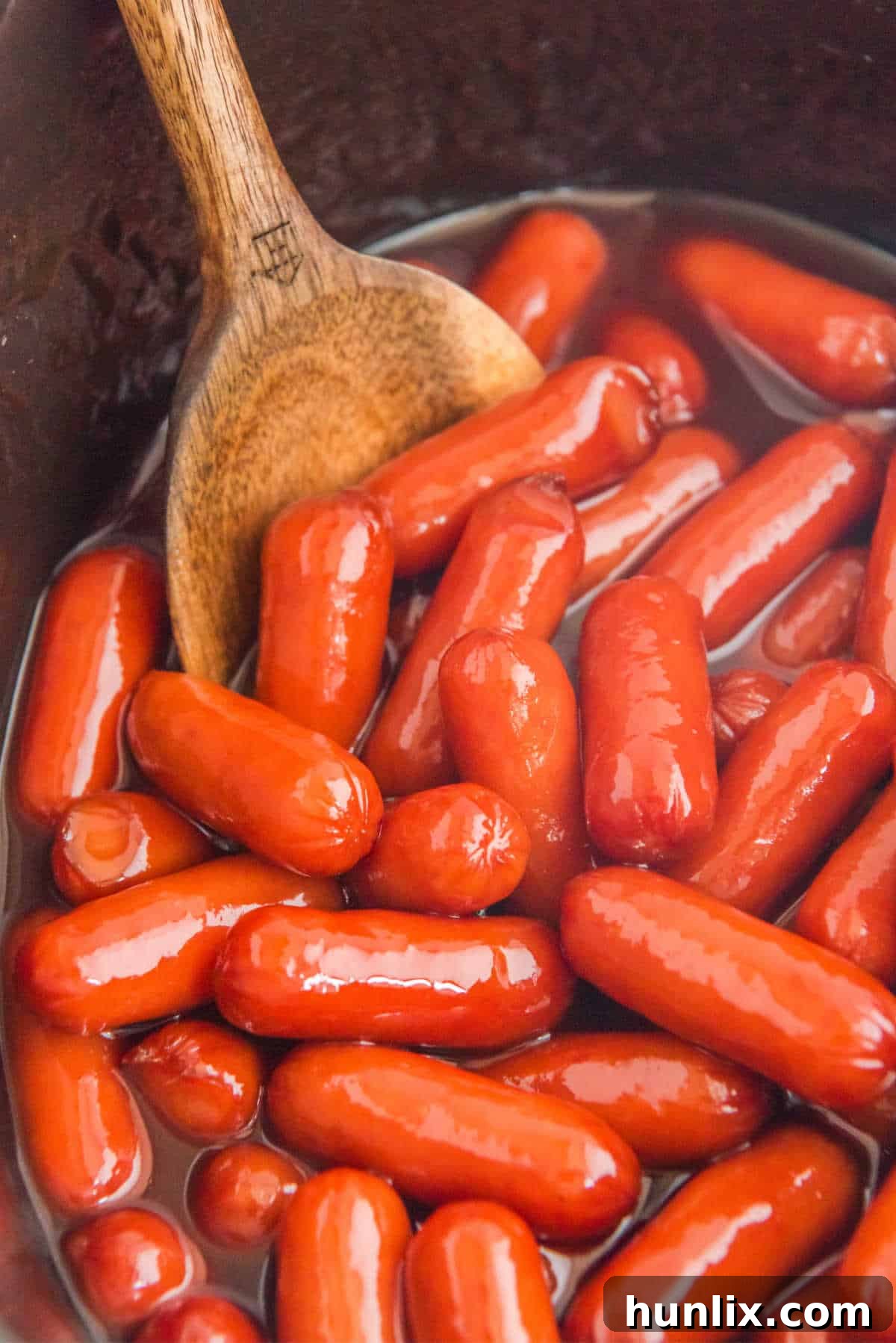 Little smokies in a slow cooker with a wooden spoon.