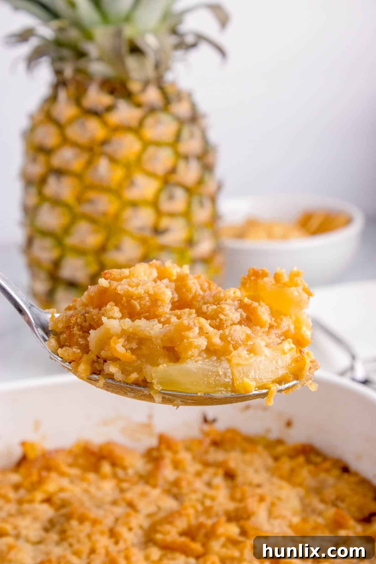 A serving spoon lifting a generous portion of pineapple casserole from a white baking dish, showing the gooey cheese, pineapple tidbits, and crispy cracker topping.
