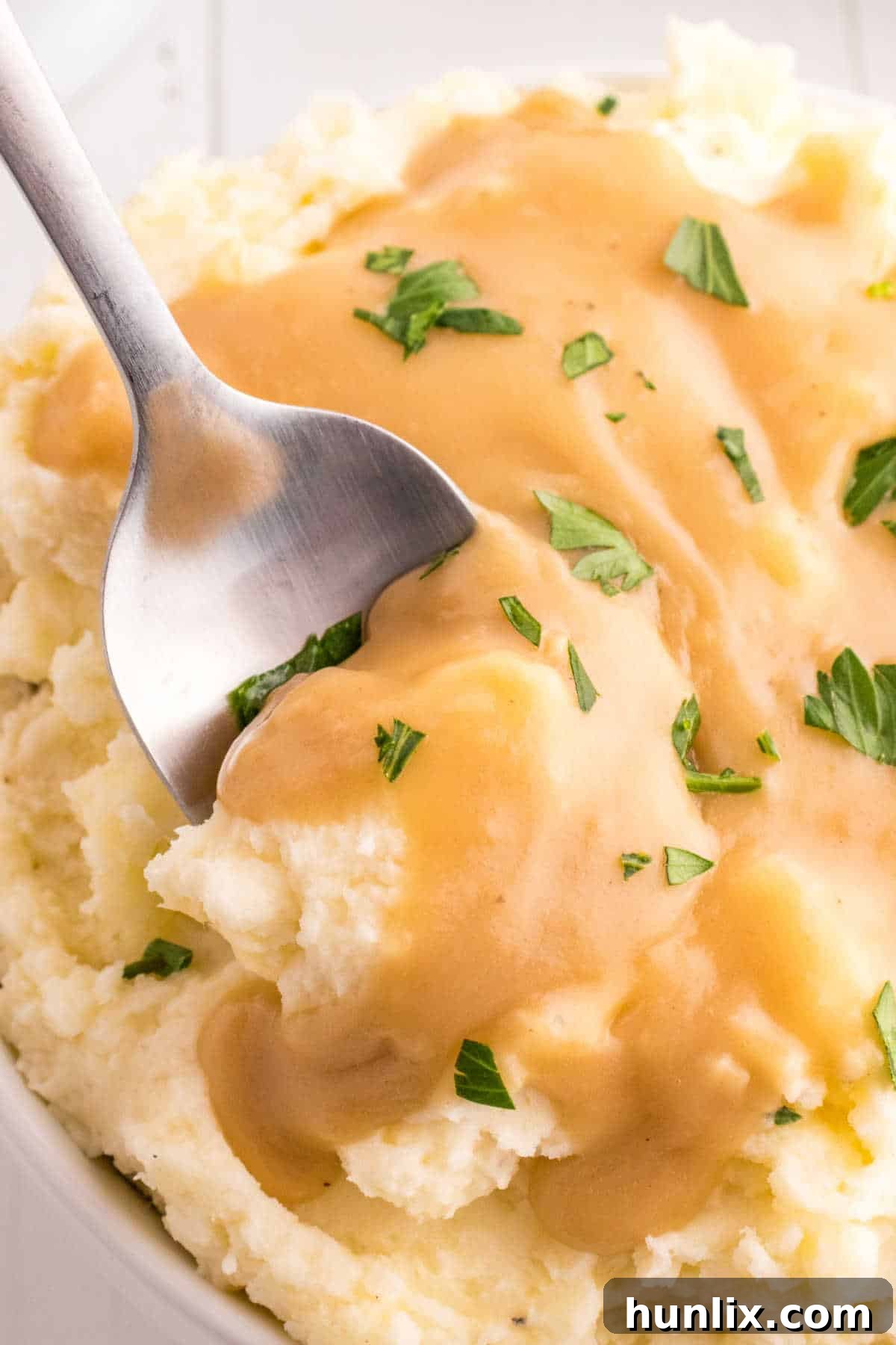 A white serving spoon resting in a bowl of creamy mashed potatoes, generously smothered with brown gravy.