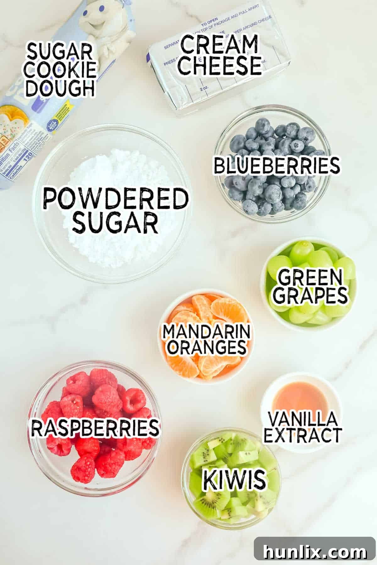 A collection of fresh ingredients laid out on a white surface, including cookie dough, cream cheese, powdered sugar, and various colorful fruits, ready to make a sugar cookie Christmas tree.