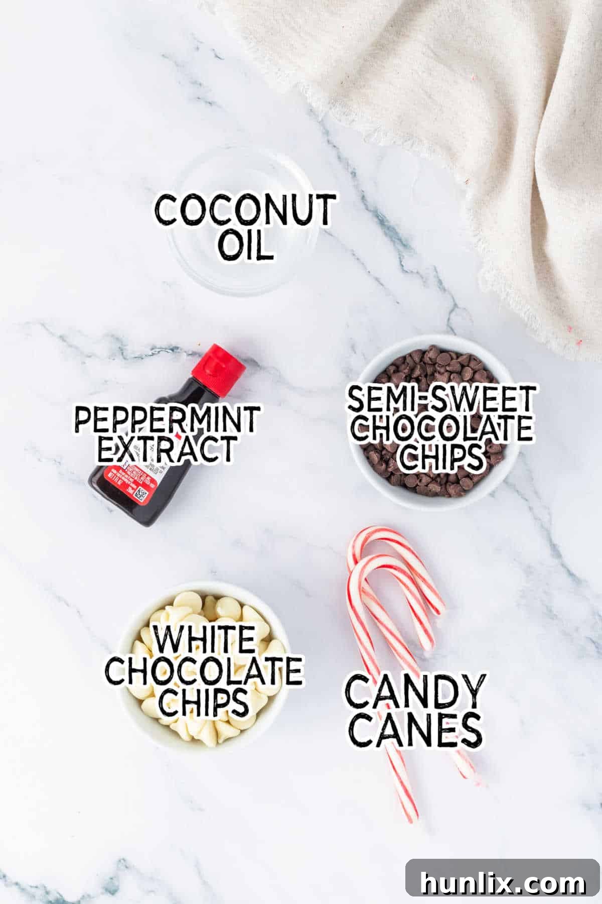 A selection of ingredients for peppermint bark laid out on a marble cutting board, including white chocolate, dark chocolate, coconut oil, peppermint extract, and candy canes.