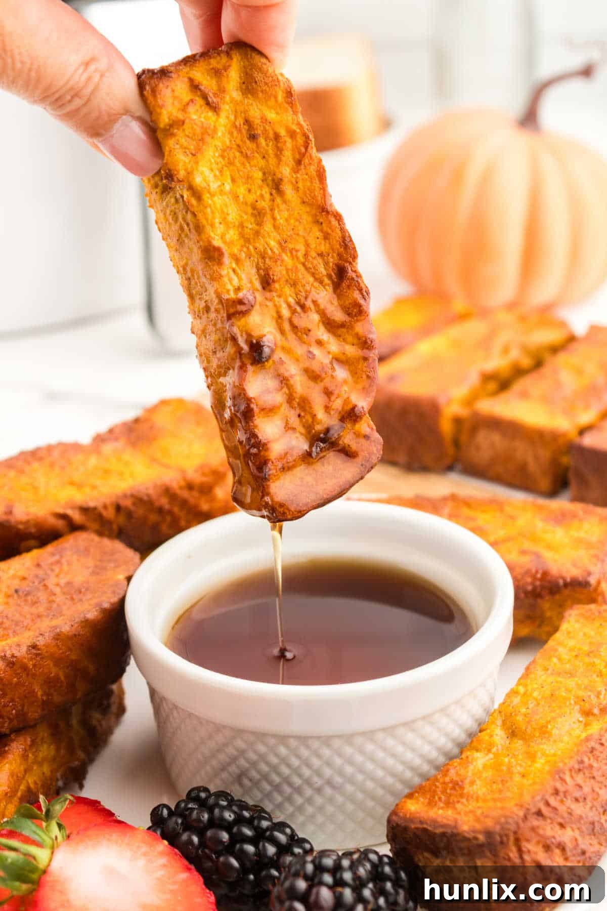 A hand dipping a perfectly golden Air Fryer Pumpkin French Toast Stick into a bowl of rich maple syrup.