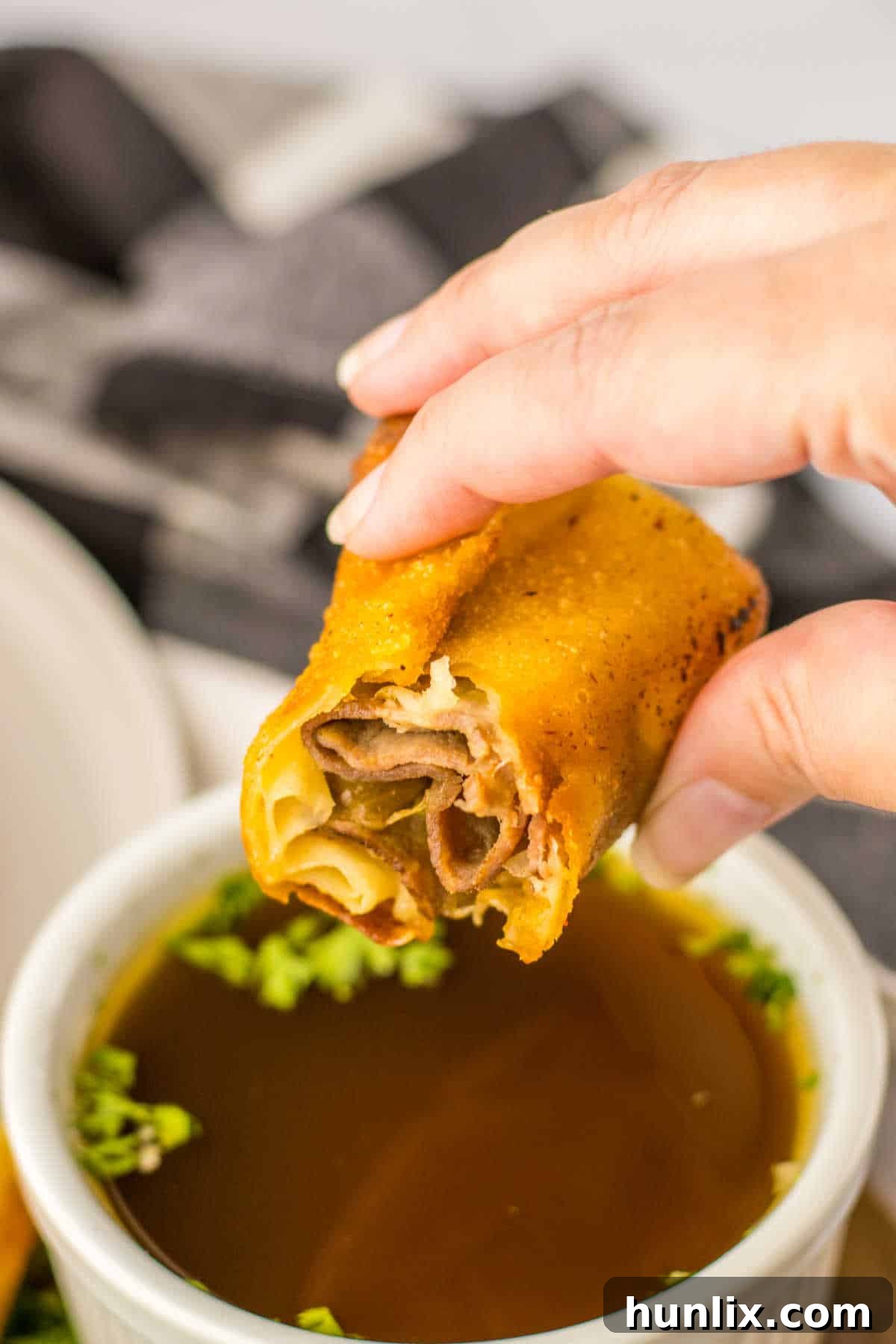Crispy French Dip Dippers 8 A hand holding a French dip egg roll cut in half, expertly dipped into a small white bowl of rich au jus sauce.