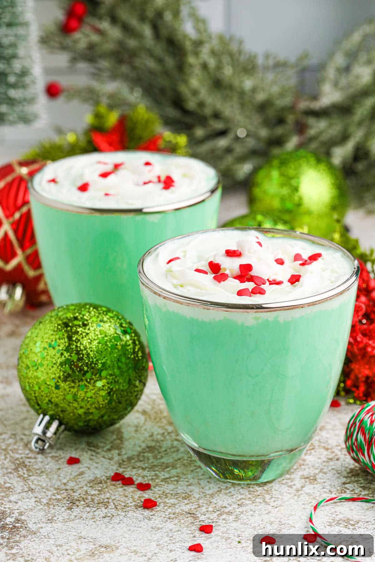 A vibrant green Grinch Mint Cocktail served in clear glass mugs, garnished with whipped cream and a red heart sprinkle, perfect for Christmas celebrations.
