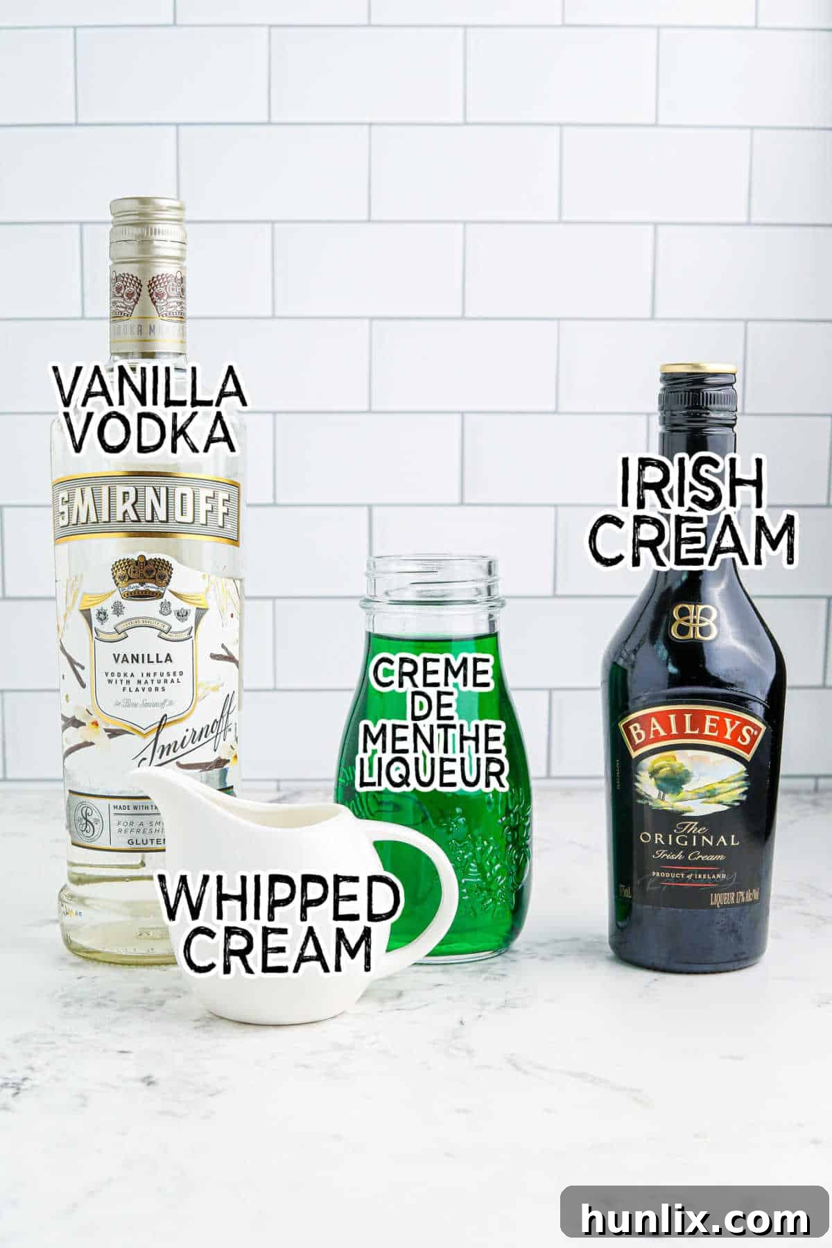 A flat lay image displaying all the ingredients for the Grinch Mint Cocktail laid out on a marble countertop, including bottles of creme de menthe, Irish cream, vanilla vodka, a bowl of ice, sprinkles, and whipped cream.