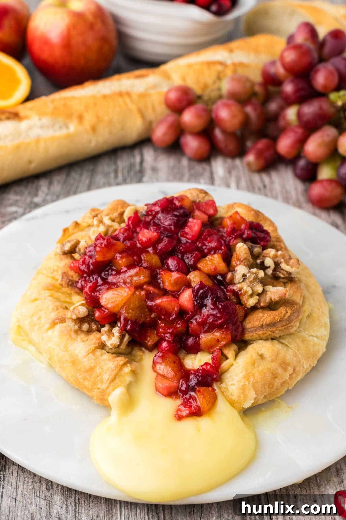 An overhead shot of baked brie with cranberry chutney and walnuts, surrounded by fresh fruit slices and grapes, presented on a white plate with a knife for serving.