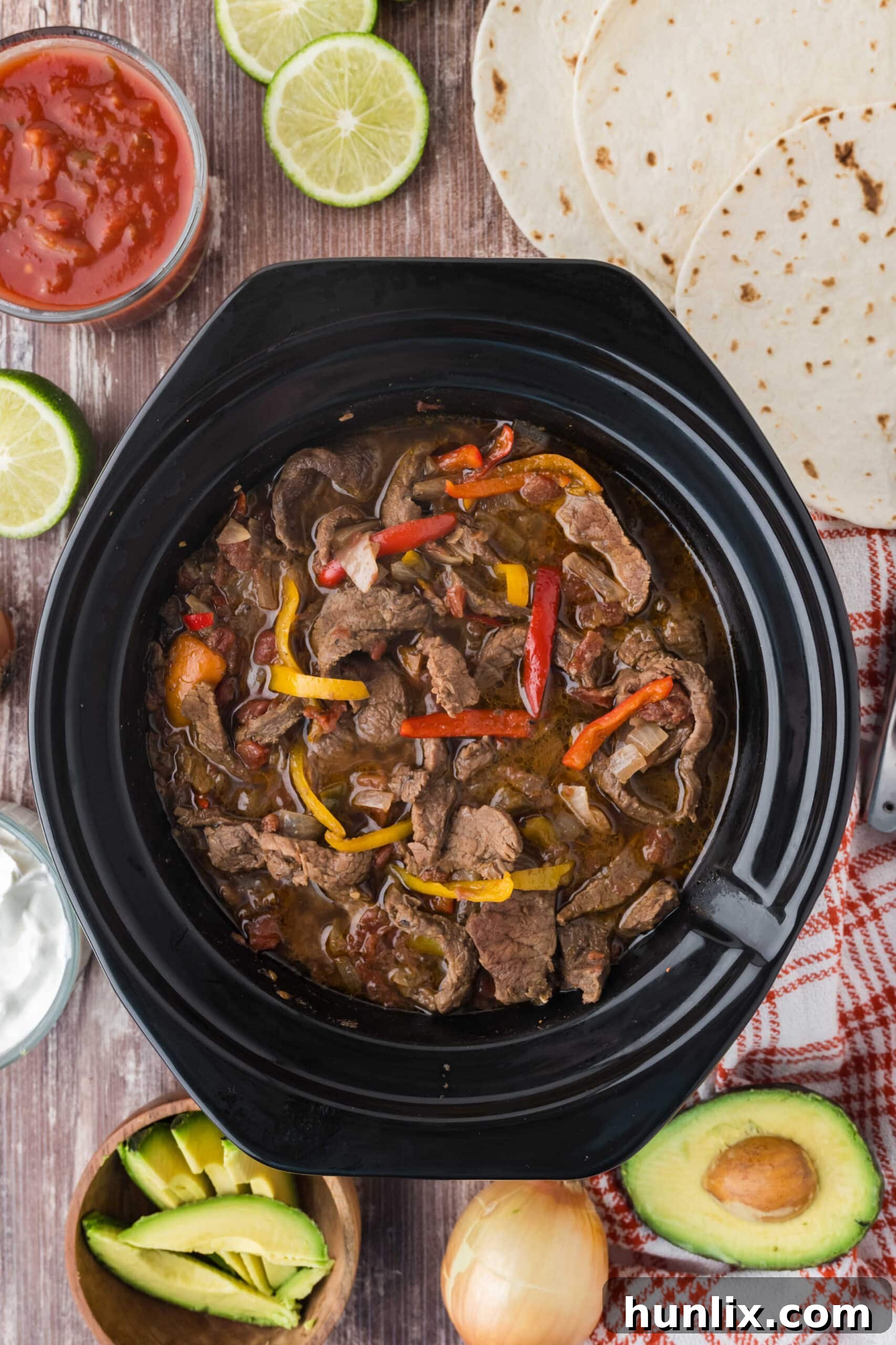 Steak fajitas cooking in a slow cooker, surrounded by fresh toppings like lime and cilantro.