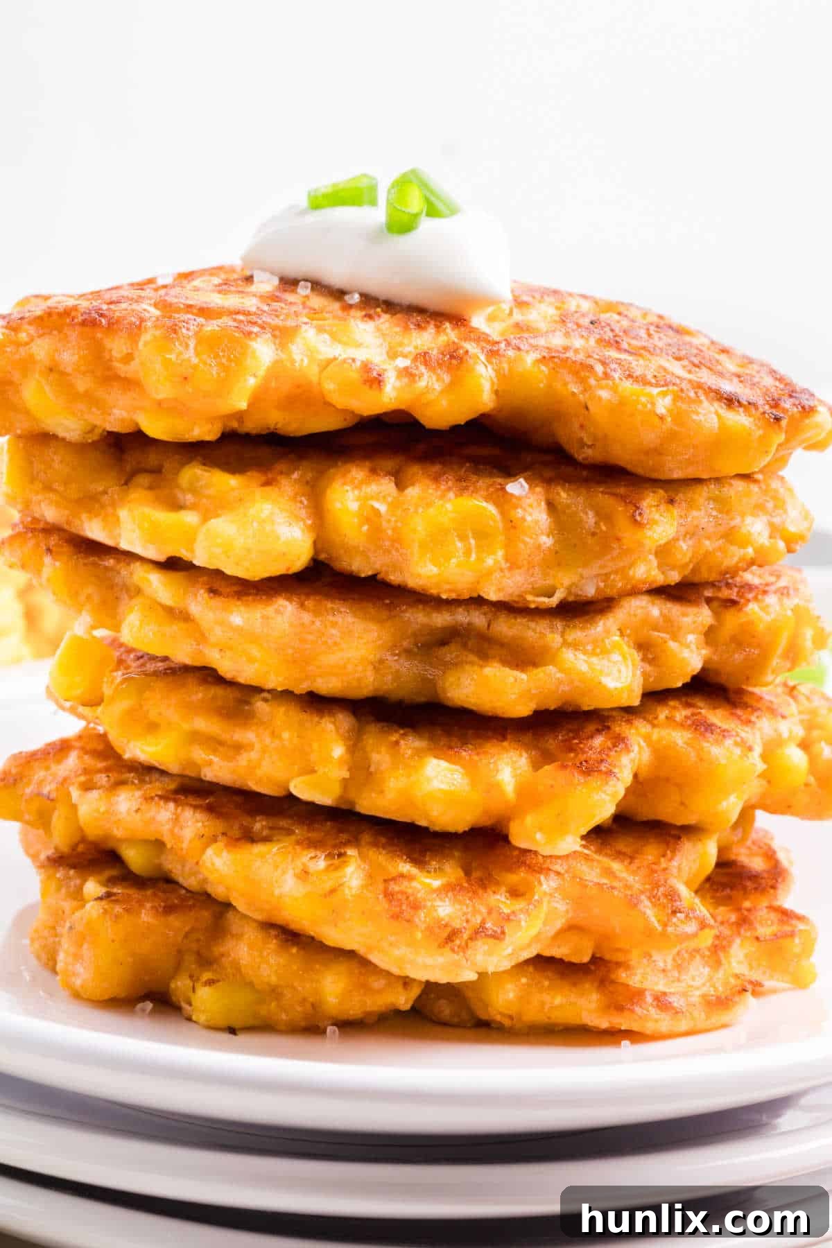 Golden Corn Bites 6 A beautifully arranged stack of golden corn fritters on a clean white plate, garnished with fresh green onions and a dollop of sour cream.