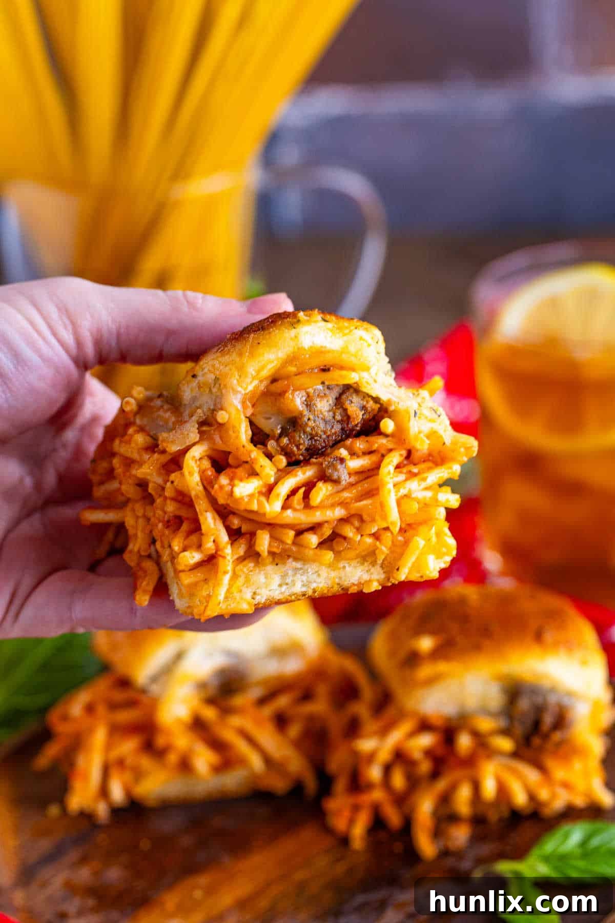 A hand holding a perfectly assembled spaghetti and meatball slider, showing the layers of pasta, meatball, and cheese.