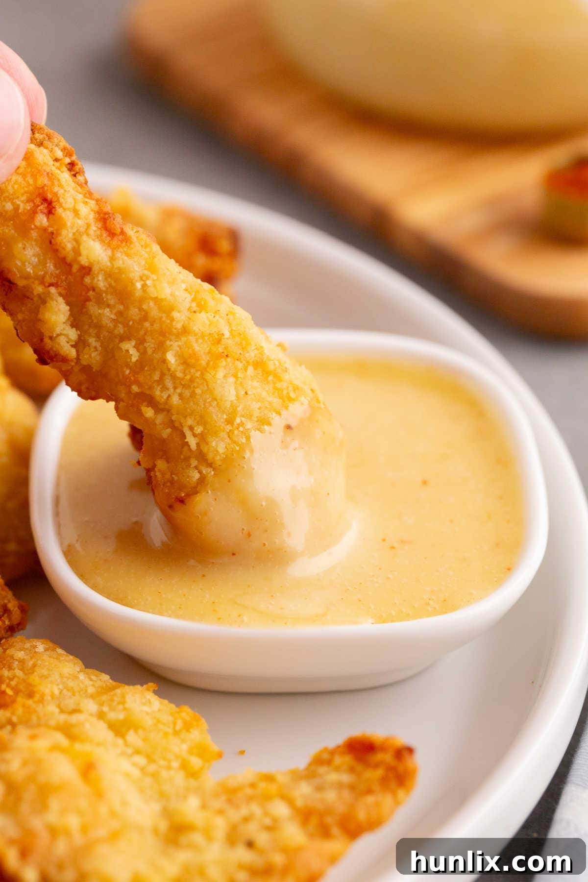 Golden Honey Mustard 2 A crispy chicken finger being dipped into a small dish of creamy, golden honey mustard sauce, illustrating its perfect consistency for dipping.