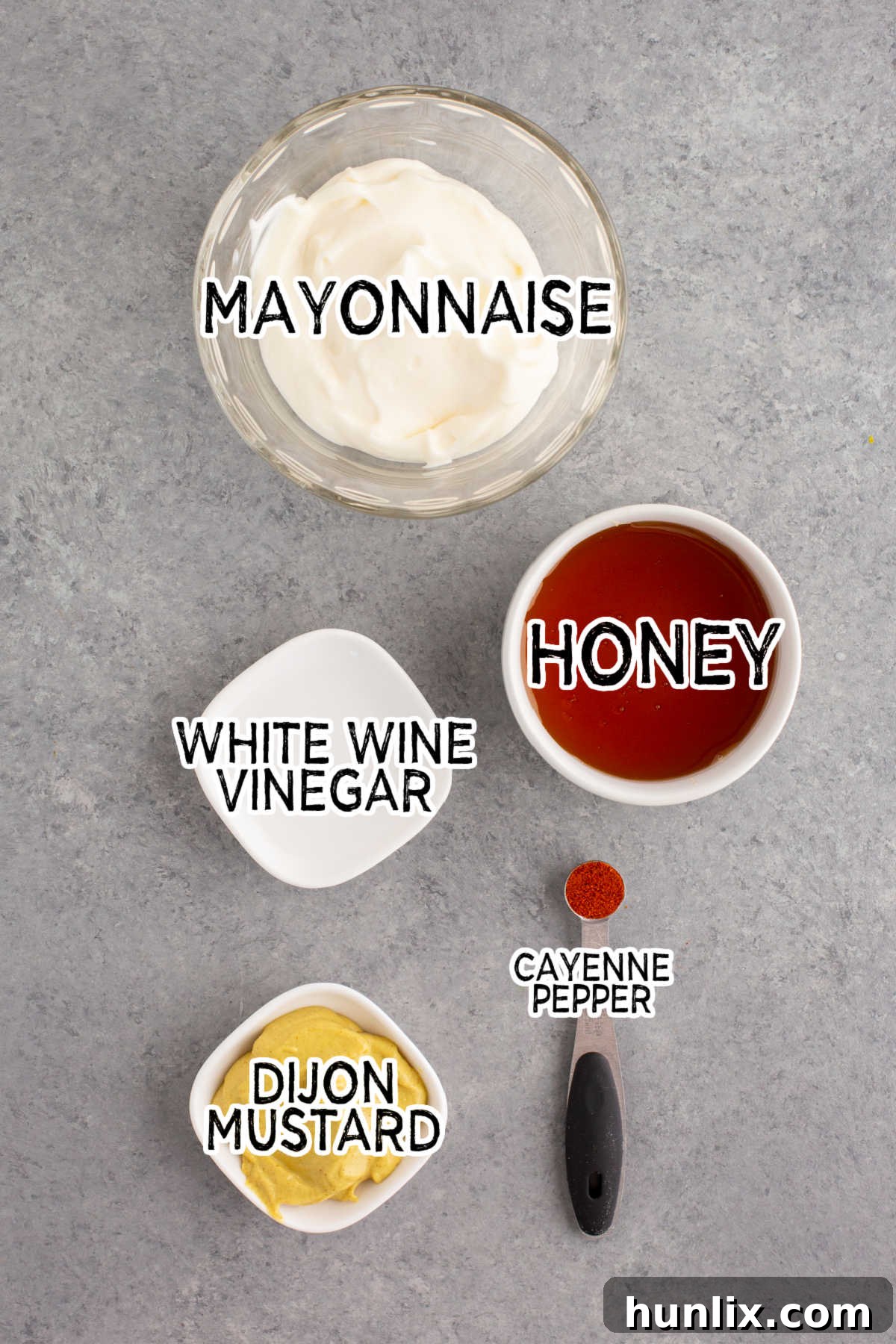 Golden Honey Mustard 3 Five ingredients for honey mustard laid out on a grey surface: a jar of mayonnaise, a bottle of honey, a bottle of Dijon mustard, a bottle of white wine vinegar, and a small container of cayenne pepper.