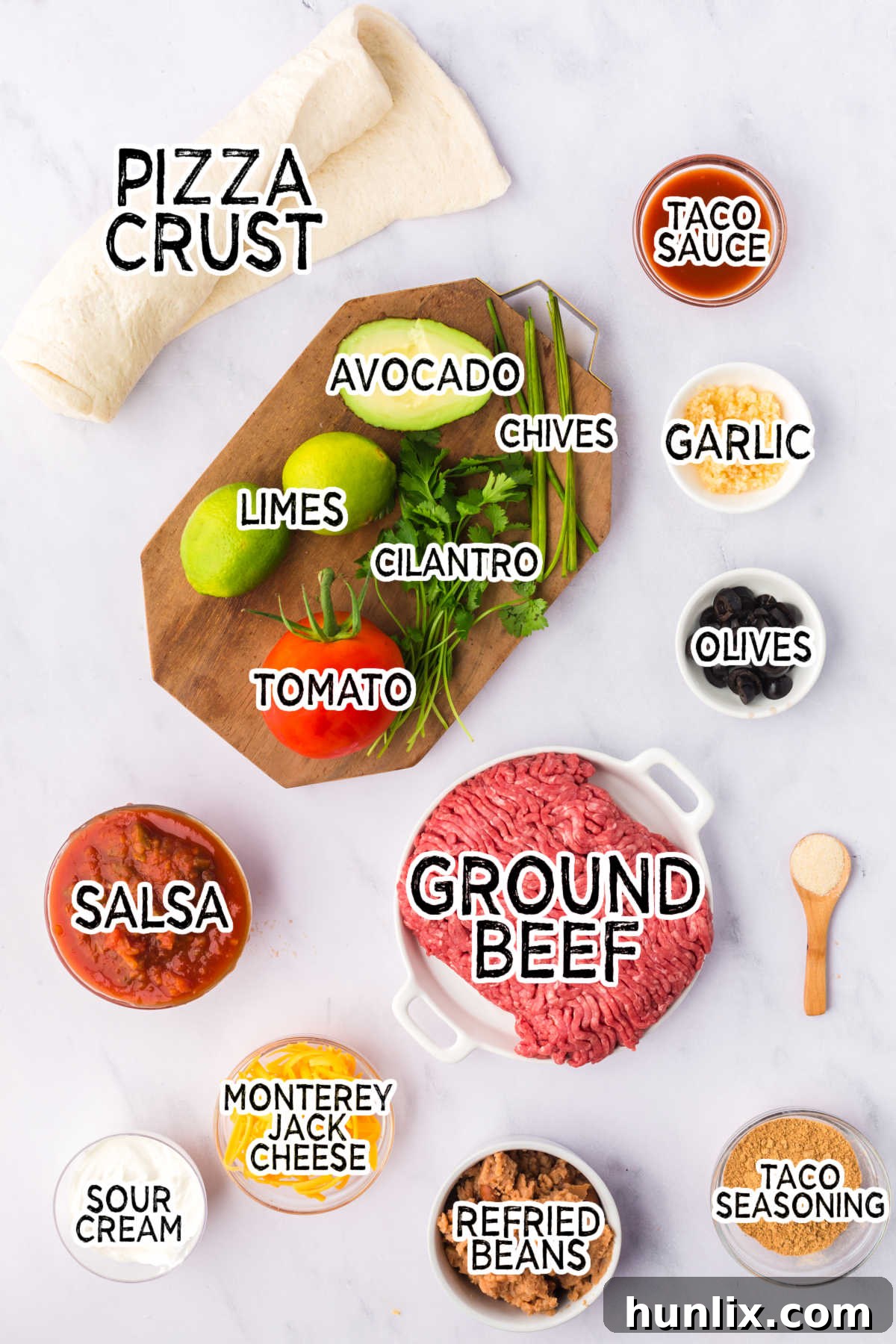 Individual ingredients for Taco Pie laid out on a clean white kitchen counter, including ground beef, pizza dough, refried beans, salsa, garlic, taco seasoning, and Monterey Jack cheese.