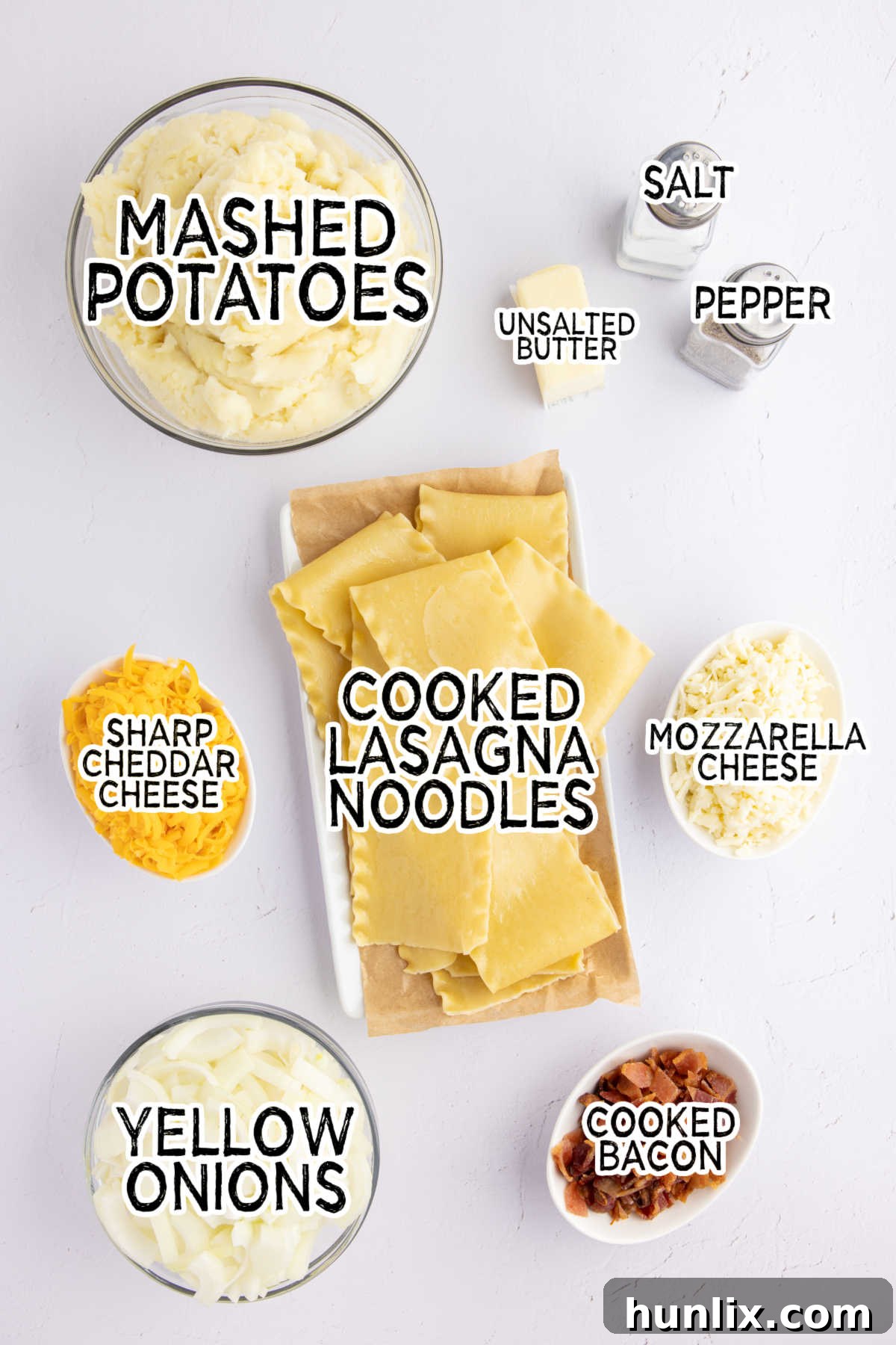 Ingredients for pierogi lasagna laid out on a white surface, including butter, onions, cheeses, noodles, potatoes, and bacon.