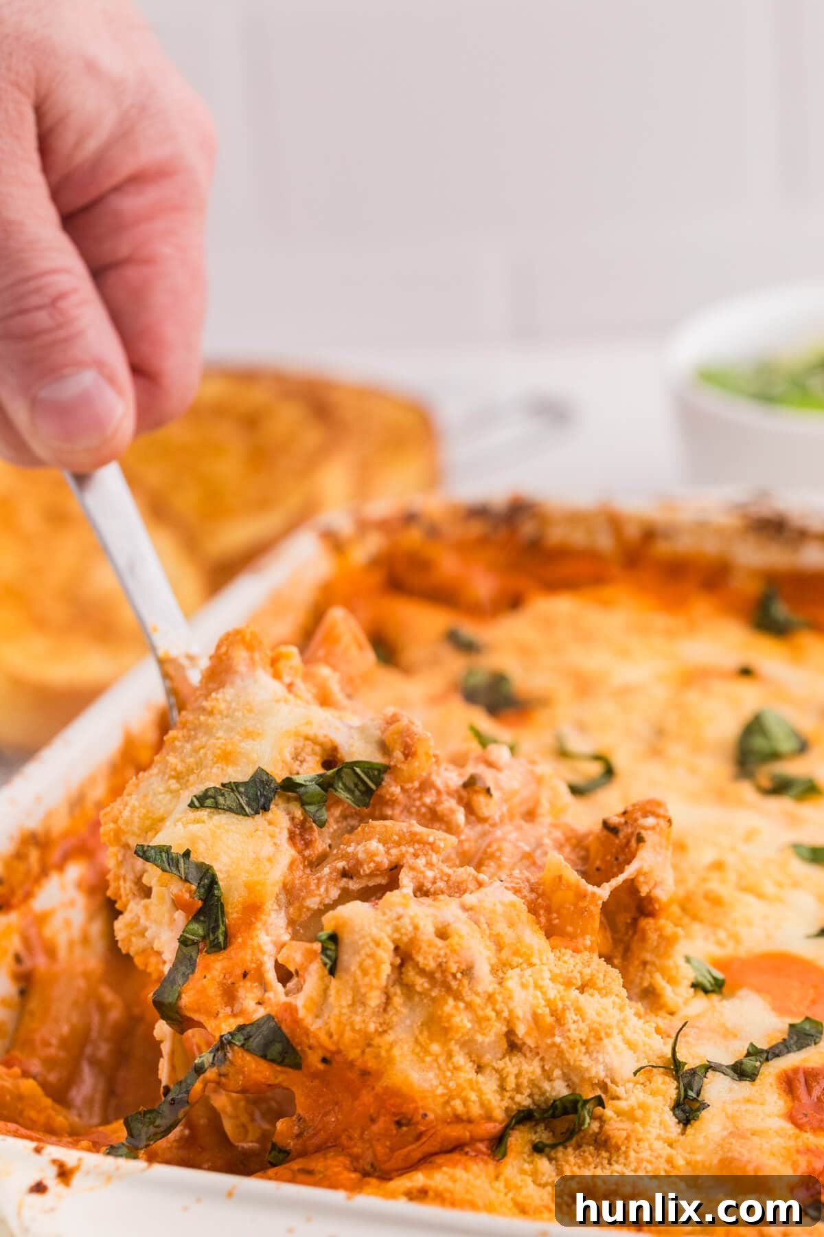 One-Dish Baked Penne 7 A hand lifting a serving spoon filled with No-Boil Baked Penne from a white casserole dish, highlighting the texture of the cooked pasta, melted cheese, and rich tomato sauce. This image emphasizes the ease of serving and the deliciousness of this one-pan comfort meal.