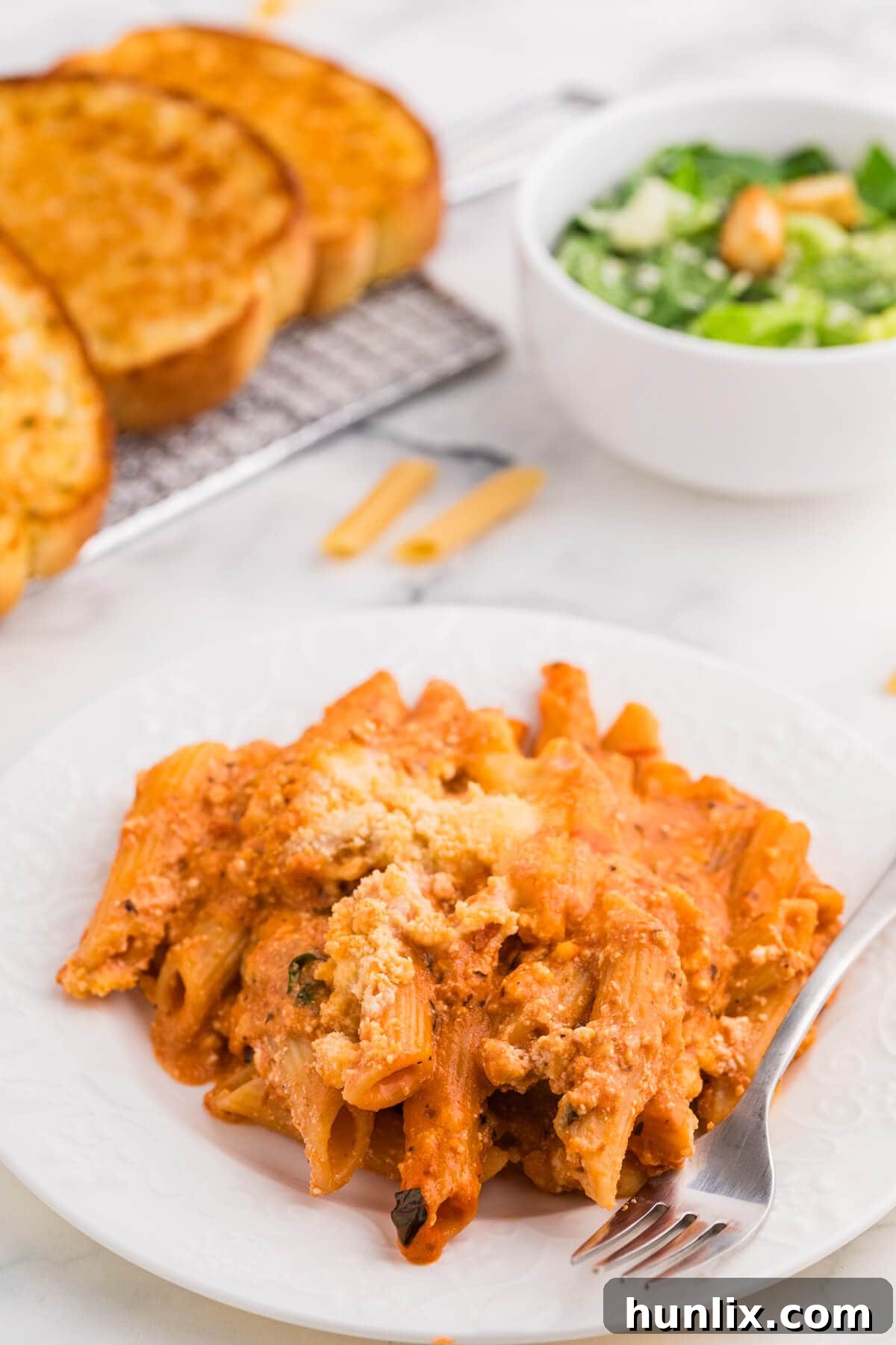 One-Dish Baked Penne 8 A single serving of No-Boil Baked Penne on a white plate, with a fork ready to take a bite, showing off the perfectly cooked penne, rich sauce, and melted cheese, emphasizing its appeal as a satisfying and easy meal.