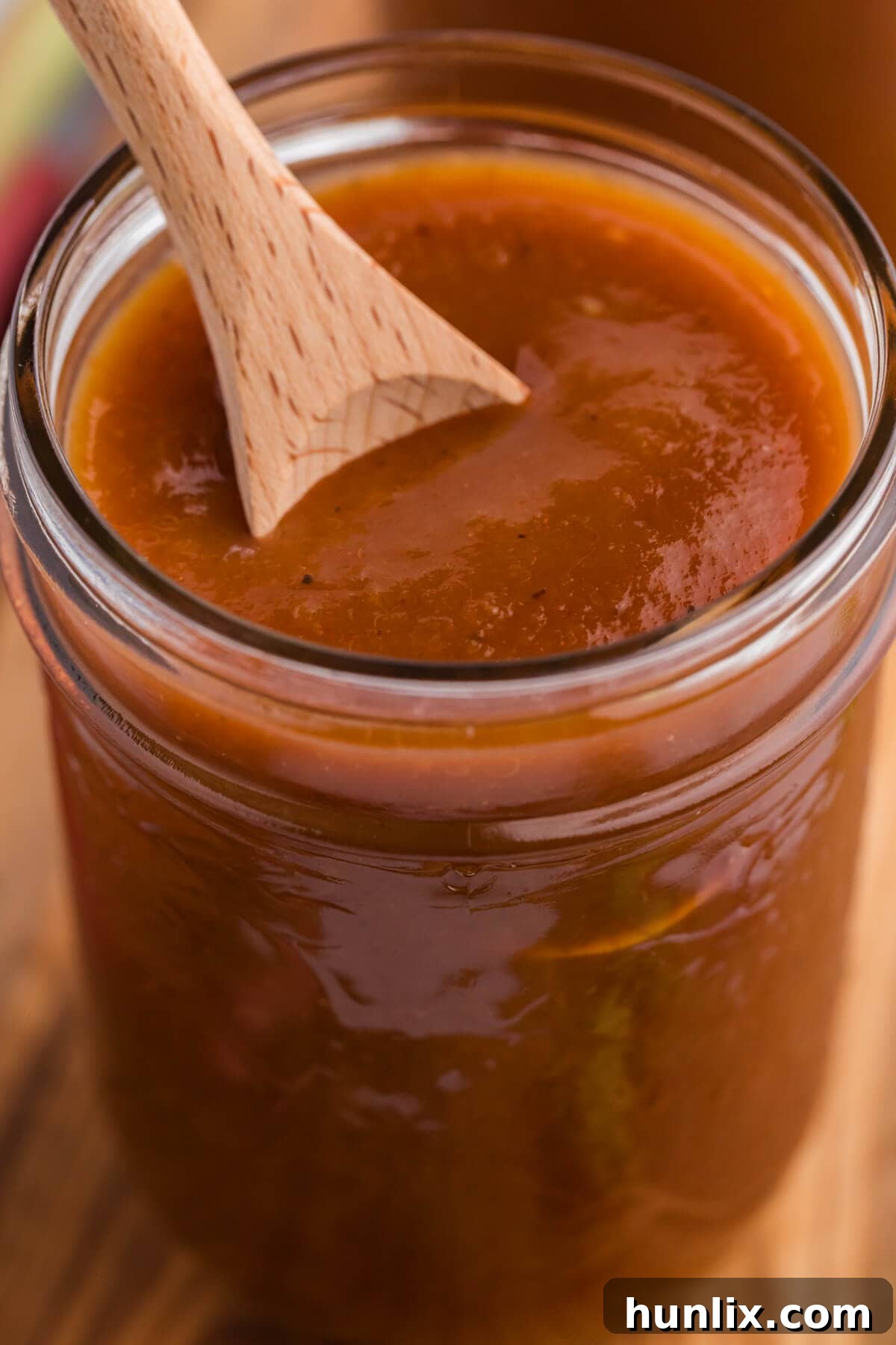 Vibrant Rhubarb BBQ Sauce in a clear glass jar, with a wooden spoon dipped in, showcasing its rich texture and appealing color. The jar sits on a rustic wooden surface, hinting at homemade goodness.