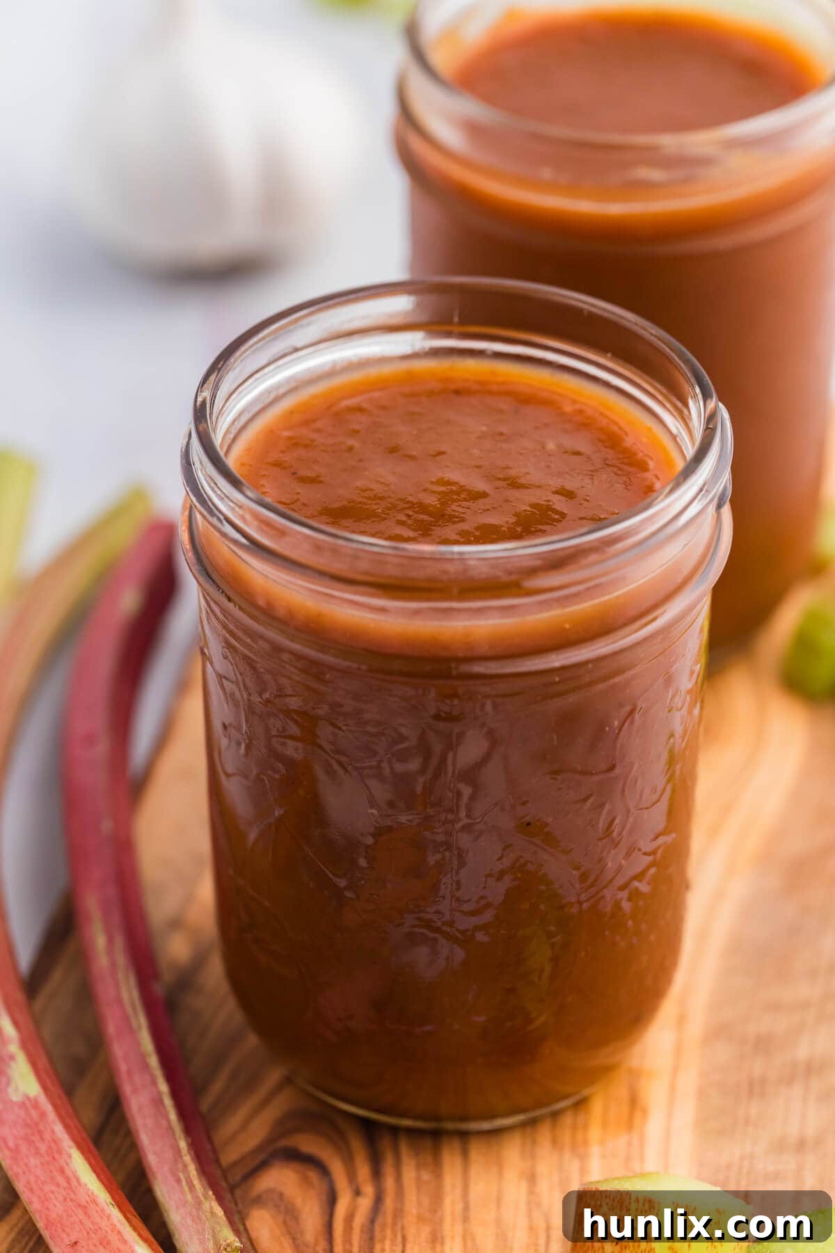 A jar of homemade Rhubarb BBQ Sauce with a wooden spoon, placed on a rustic wooden cutting board, ready to be enjoyed.