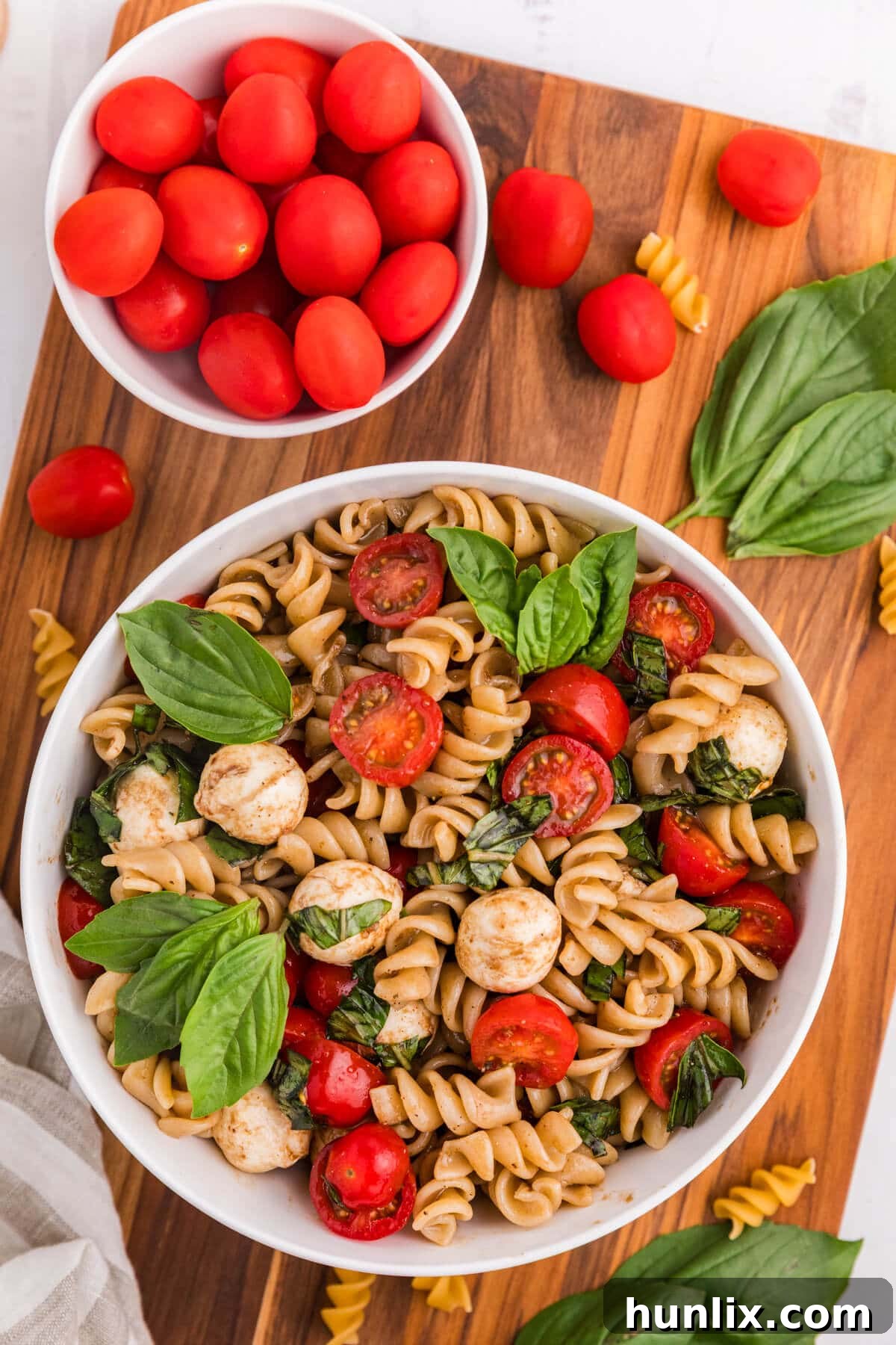 A beautiful bowl of Caprese Pasta Salad on a wooden cutting board, surrounded by fresh ingredients like tomatoes, mozzarella, and basil.