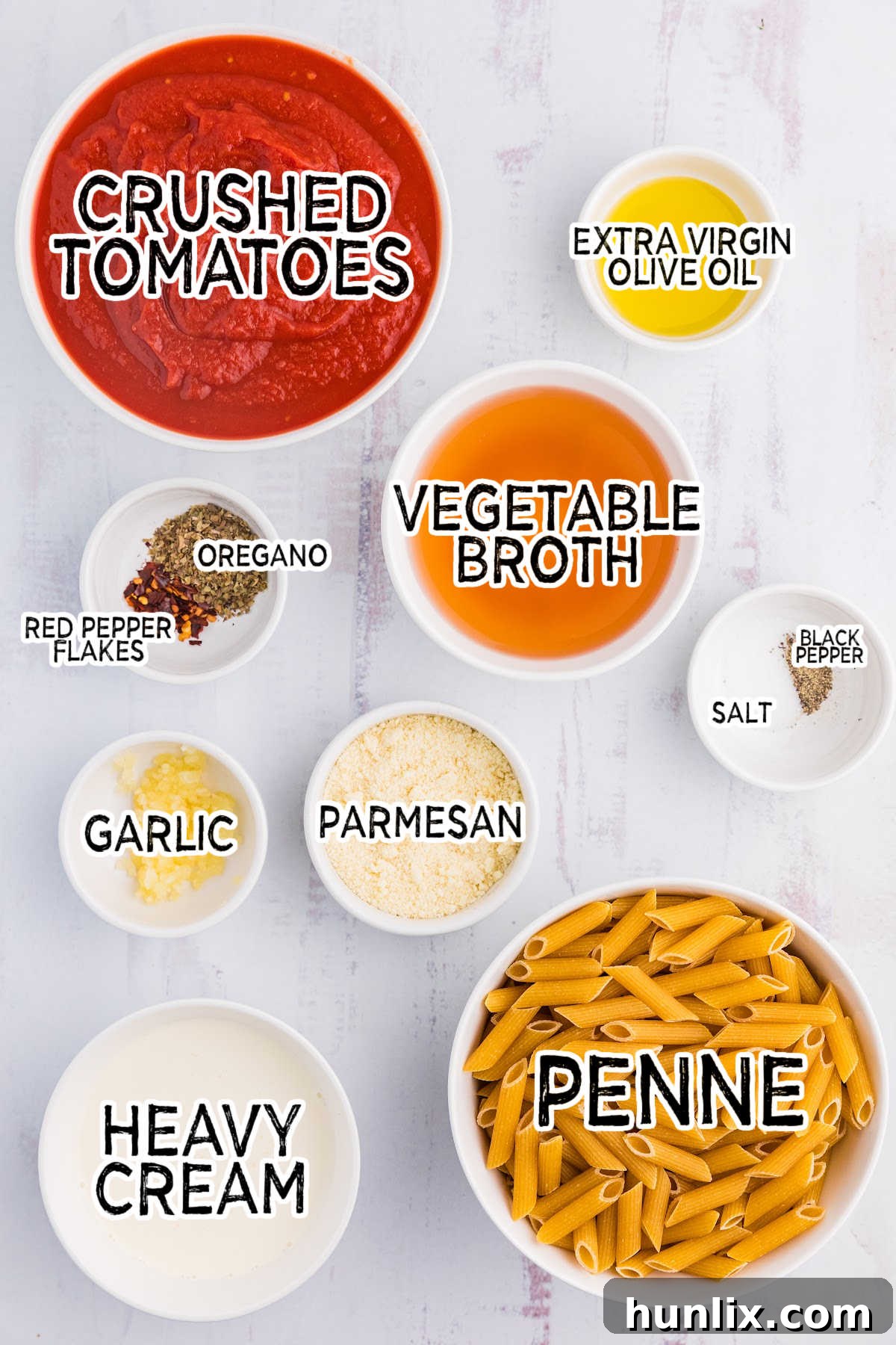 Various fresh and pantry ingredients laid out on a white background, including olive oil, garlic, oregano, red pepper flakes, canned crushed tomatoes, vegetable broth, heavy cream, penne pasta, salt, pepper, Parmesan cheese, and fresh basil.