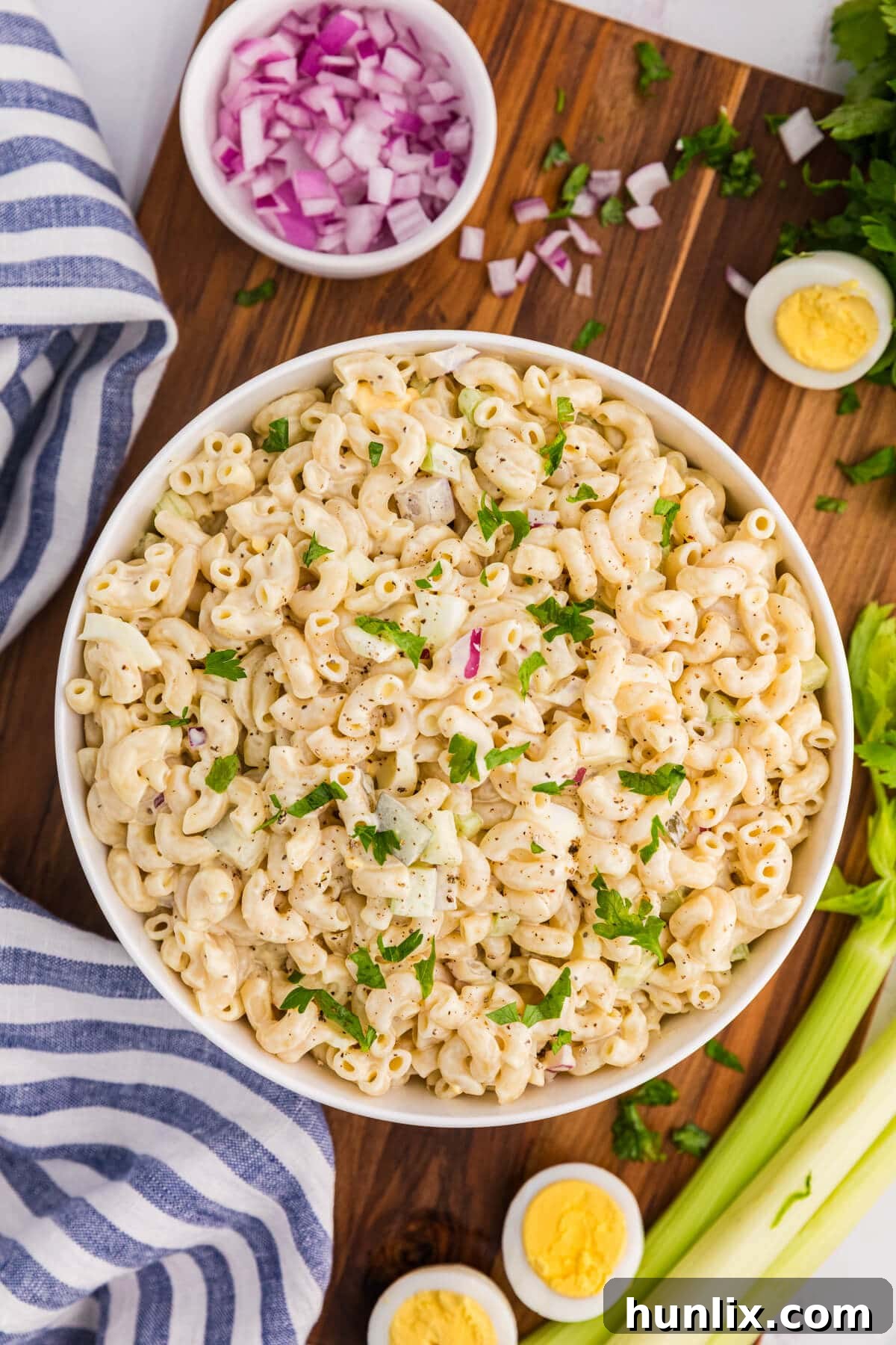 Classic creamy macaroni salad on a wooden surface, surrounded by fresh ingredients like celery, onions, and pickles.