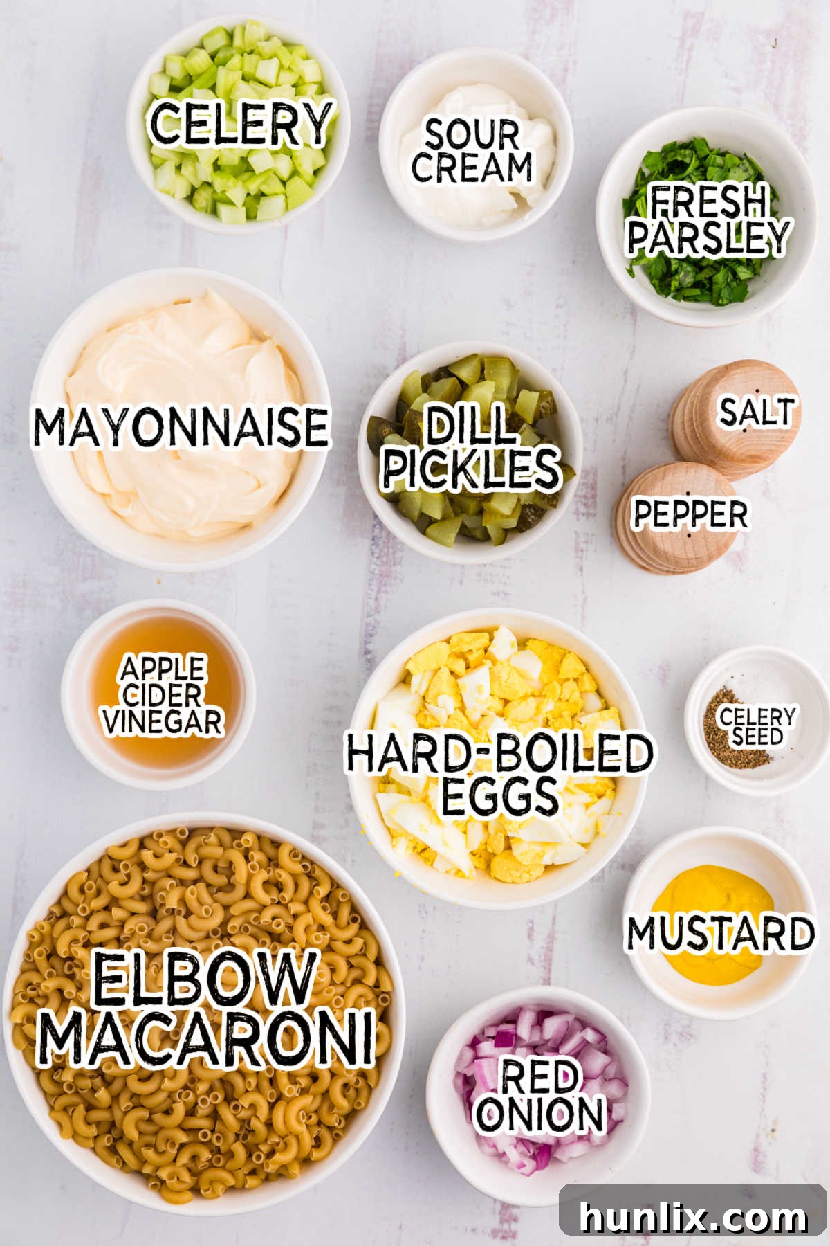 A vibrant assortment of ingredients for classic macaroni salad, including cooked elbow macaroni, chopped celery, red onion, hard-boiled eggs, and pickles, displayed on a clean white surface.