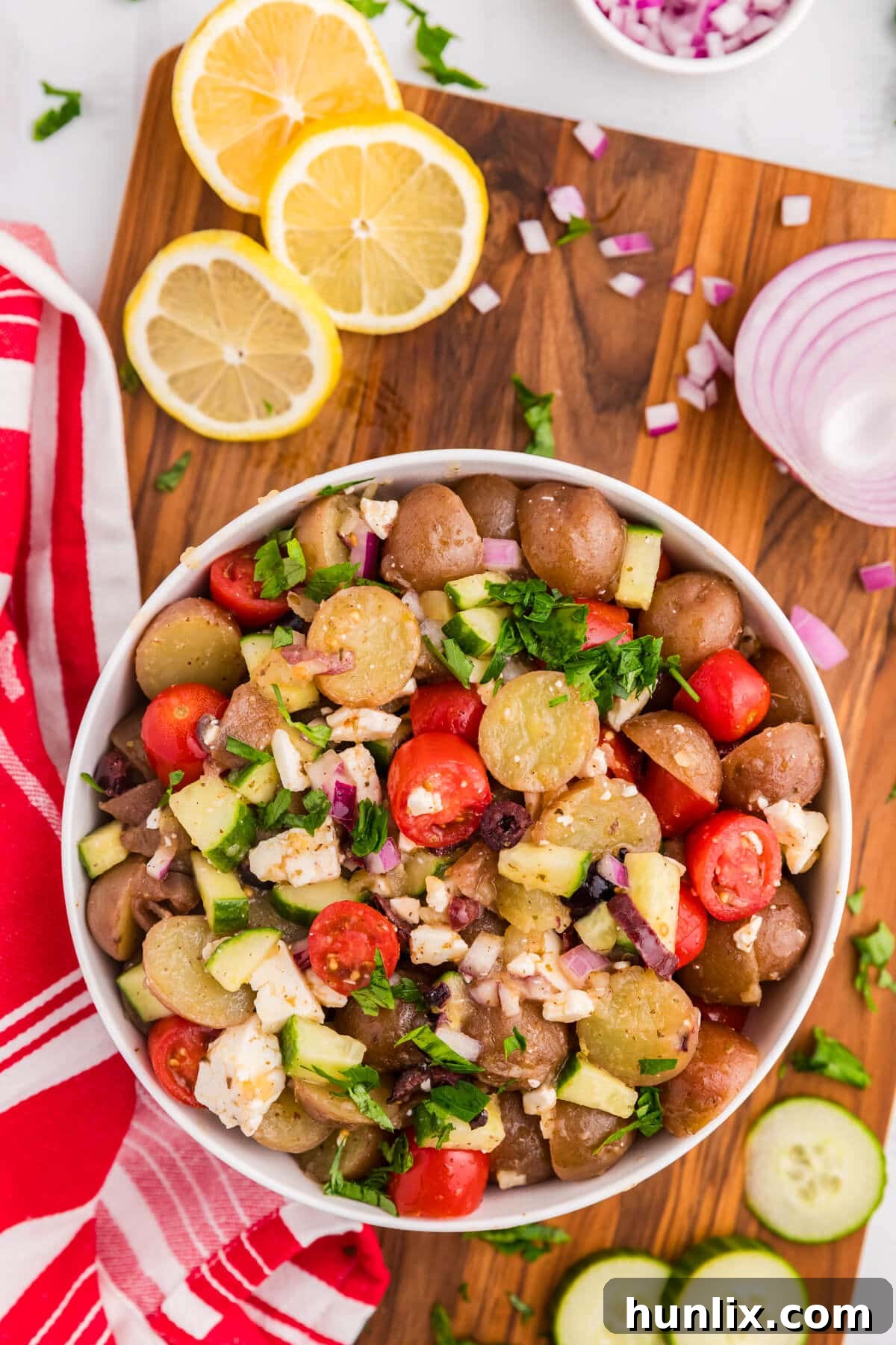 A beautifully arranged bowl of Greek Potato Salad on a rustic wooden surface, surrounded by fresh ingredients like lemons, cucumbers, and cherry tomatoes, highlighting its vibrant colors and freshness.