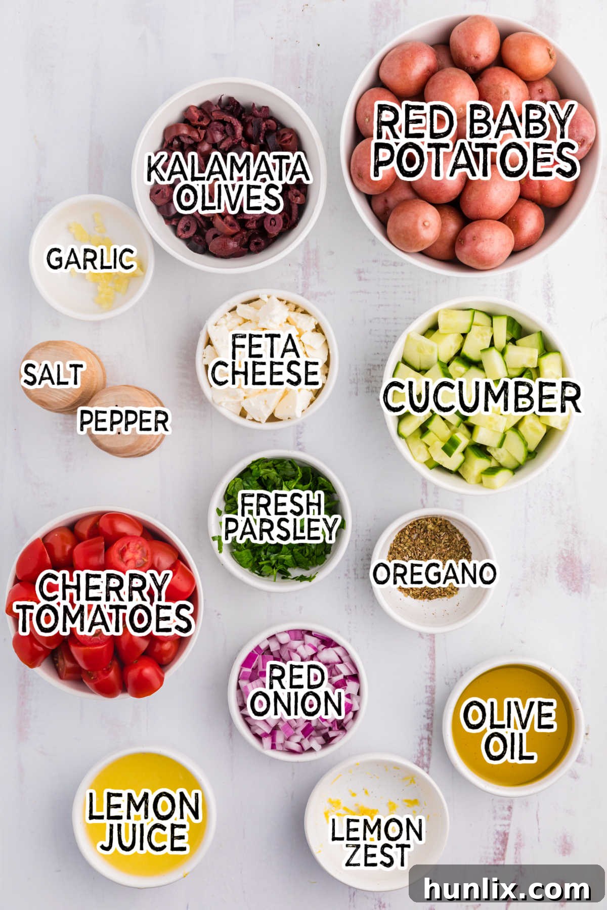 Assorted fresh ingredients for Greek Potato Salad laid out on a clean white wooden surface, including red potatoes, cucumber, cherry tomatoes, Kalamata olives, feta, red onion, lemon, olive oil, and herbs.