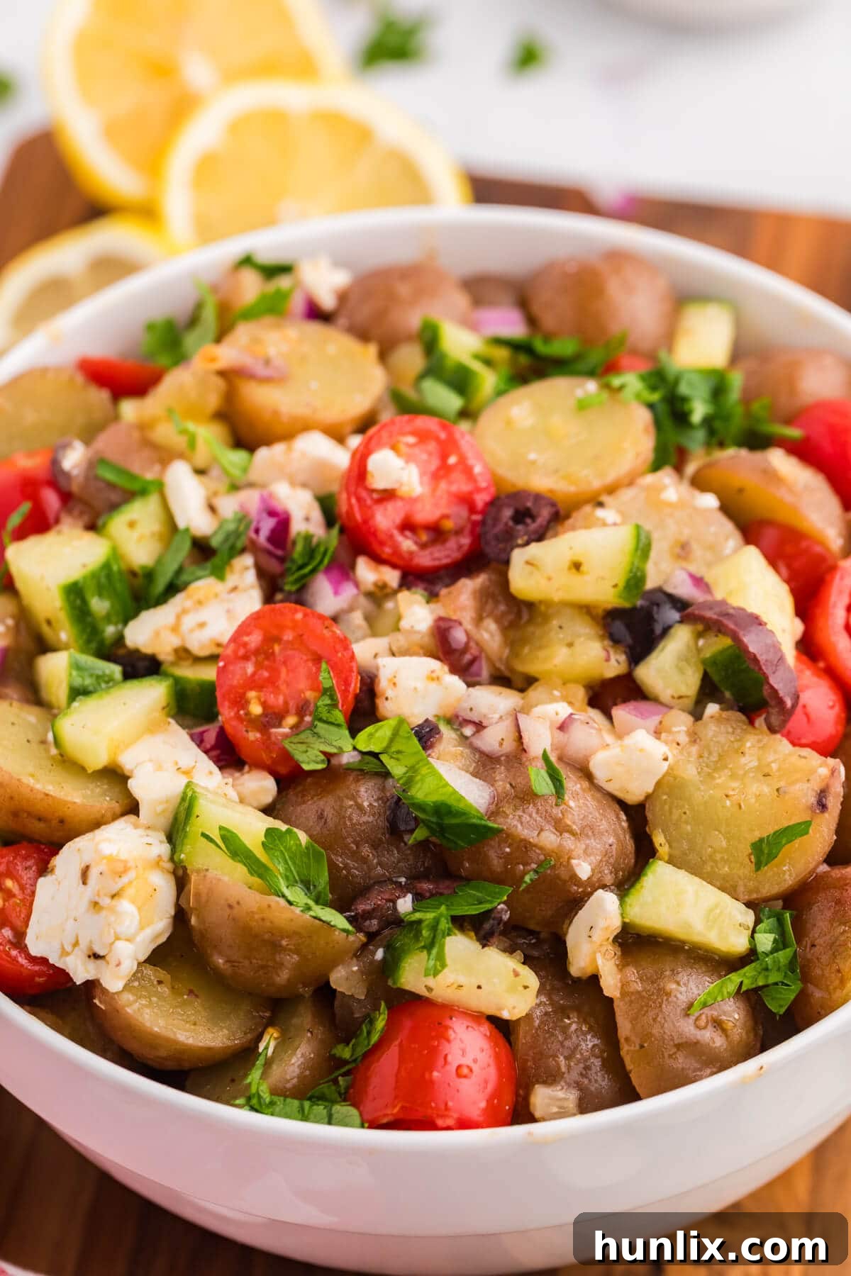 A close-up view of a serving of Greek Potato Salad in a white bowl, showcasing the vibrant ingredients like red potatoes, tomatoes, olives, and feta, with a fork ready to scoop.