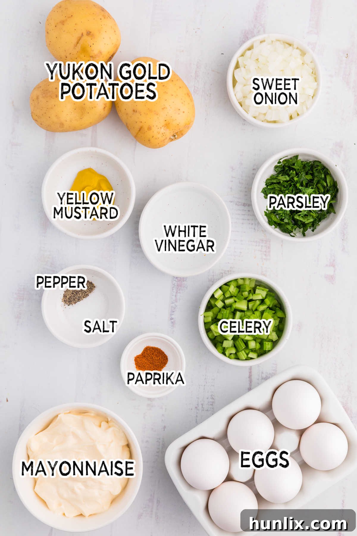 A collection of fresh ingredients for Old-Fashioned Potato Salad, including Yukon Gold potatoes, hard-boiled eggs, mayonnaise, mustard, celery, and sweet onion, arranged neatly on a clean white background.