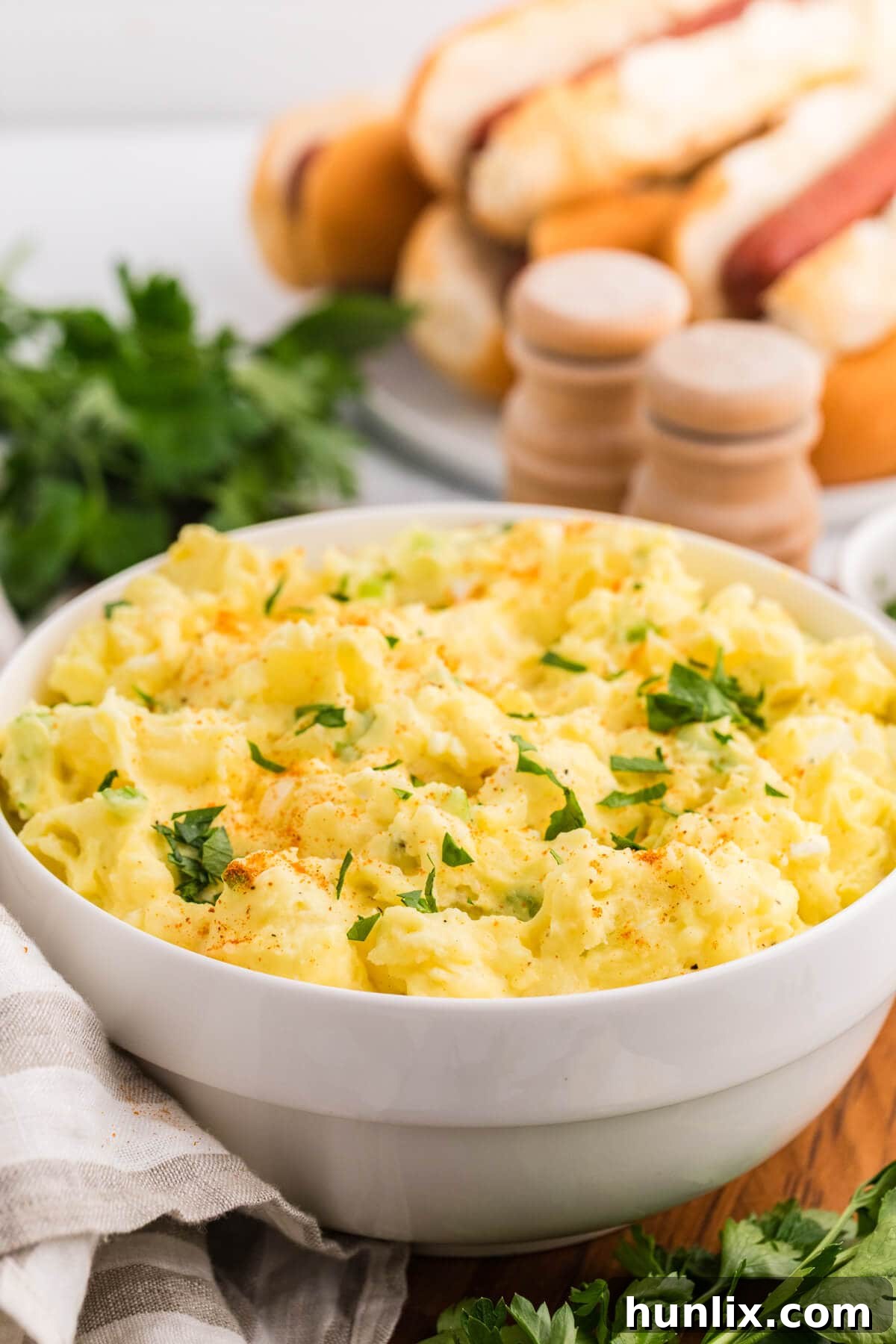 A generous bowl of Old-Fashioned Potato Salad, expertly garnished and ready to serve, positioned on a textured wooden background. The lighting highlights its creamy texture and fresh ingredients.