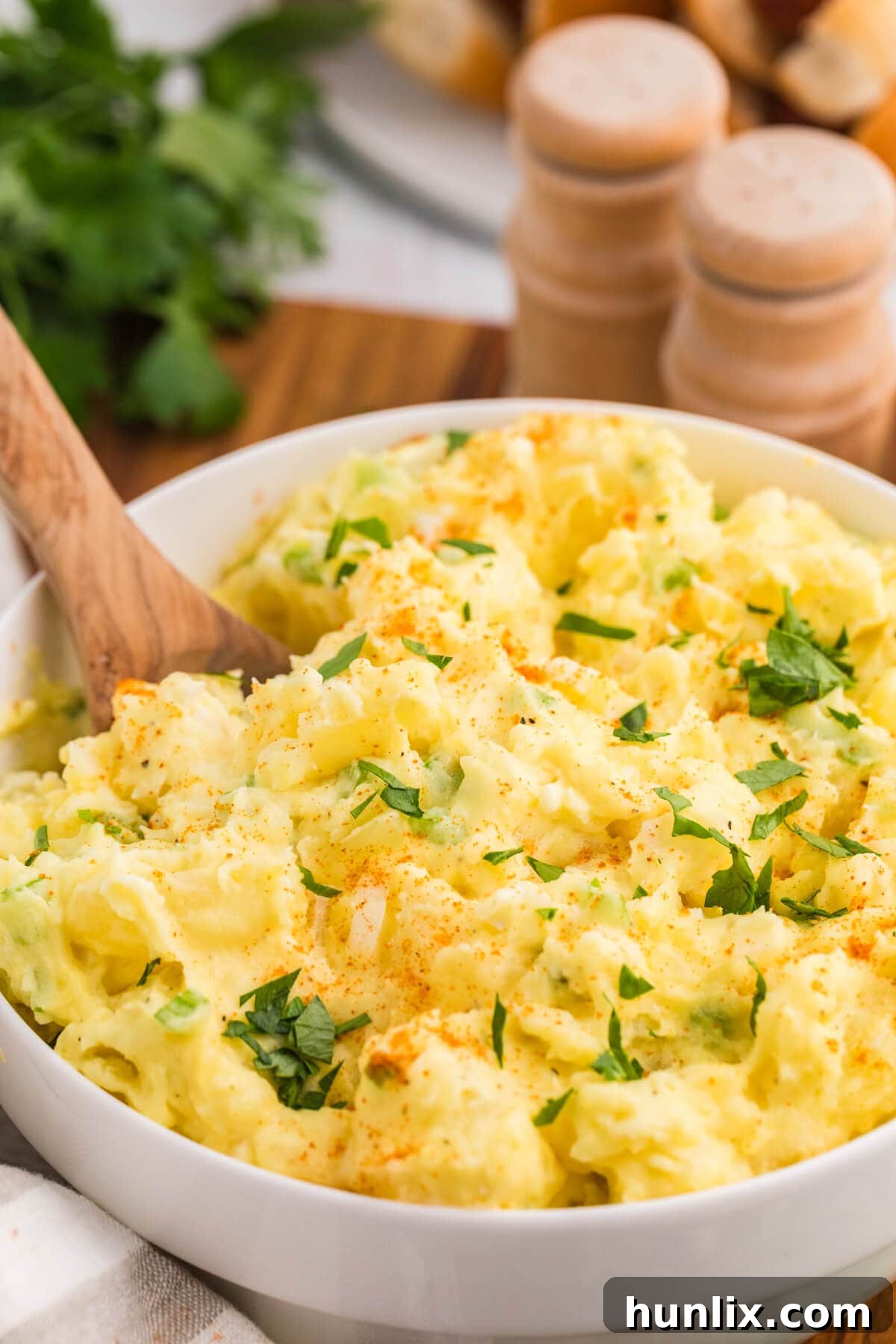 A single serving of Old-Fashioned Potato Salad scooped onto a wooden spoon, showcasing its creamy texture and the vibrant specks of celery and paprika. The full bowl is visible in the soft-focused background.