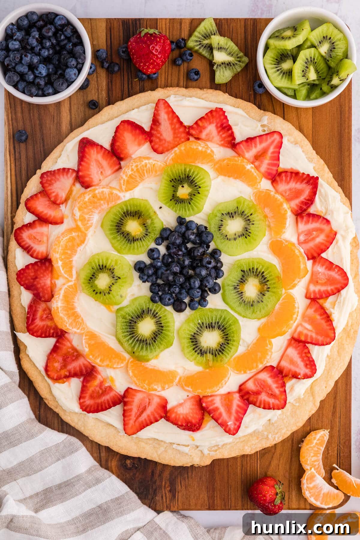 A beautifully arranged fruit pizza on a rustic wooden surface, surrounded by fresh fruits like strawberries, blueberries, and kiwi slices, highlighting its vibrant colors and inviting texture.