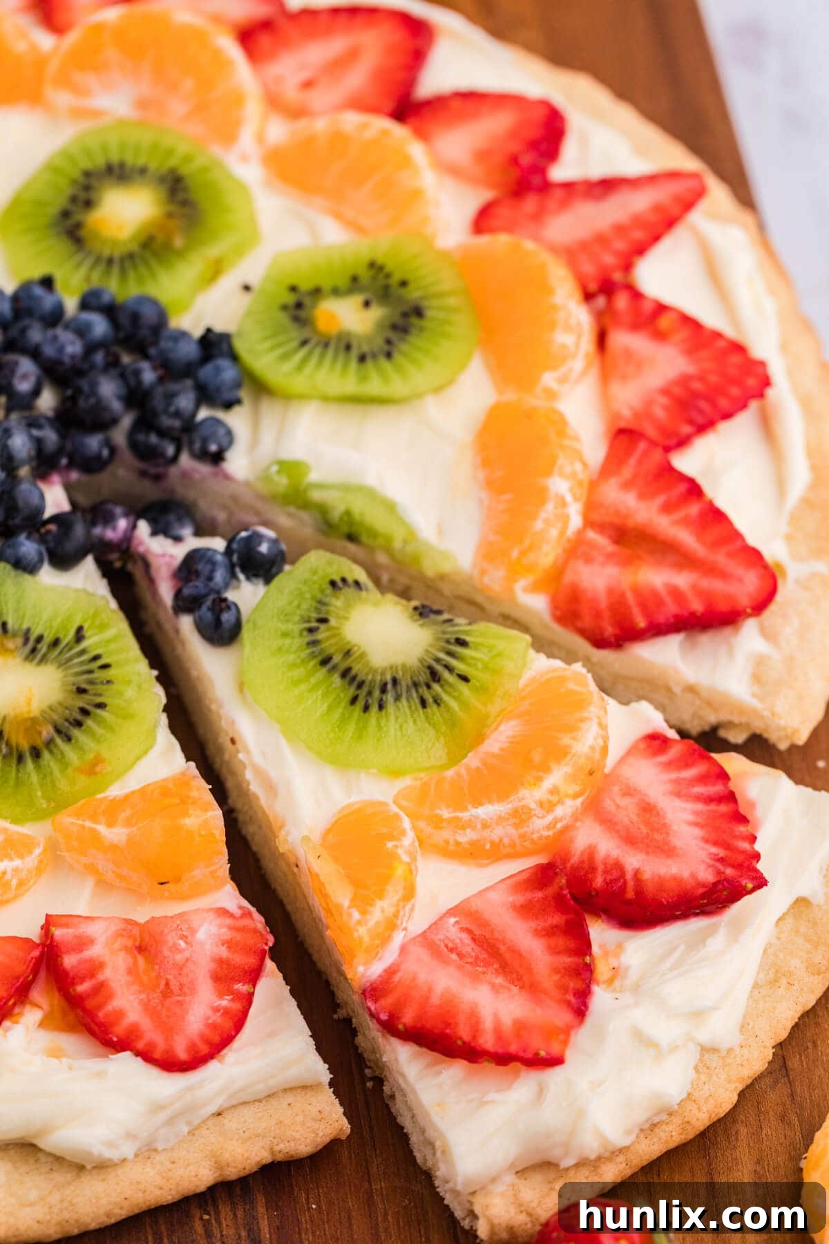 A single slice of fruit pizza, perfectly cut to show the layers of golden crust, creamy white frosting, and colorful fresh fruit, highlighted on a wooden background.