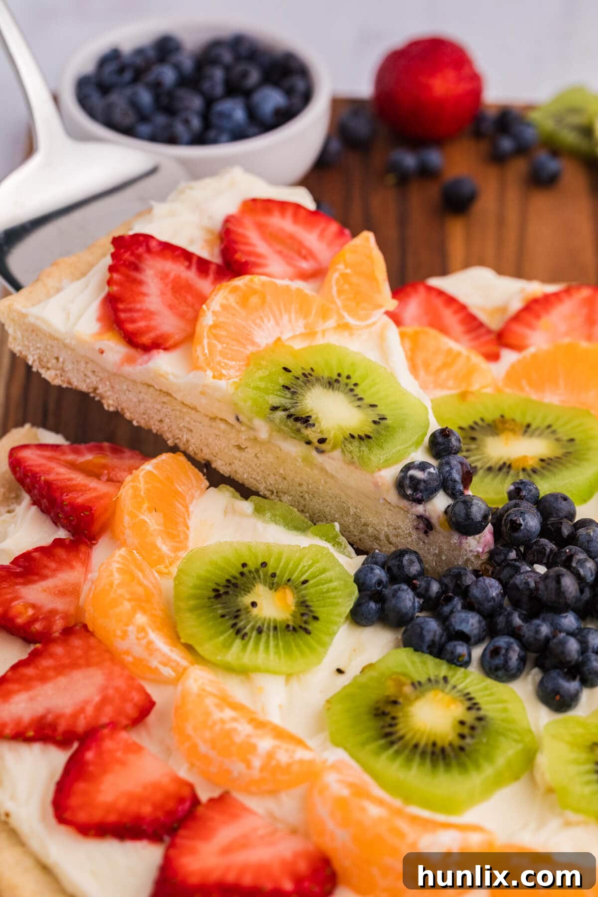 A perfectly cut slice of fruit pizza, showcasing its layers of crisp crust, creamy frosting, and colorful fruit, presented on a serving spatula ready to be enjoyed.