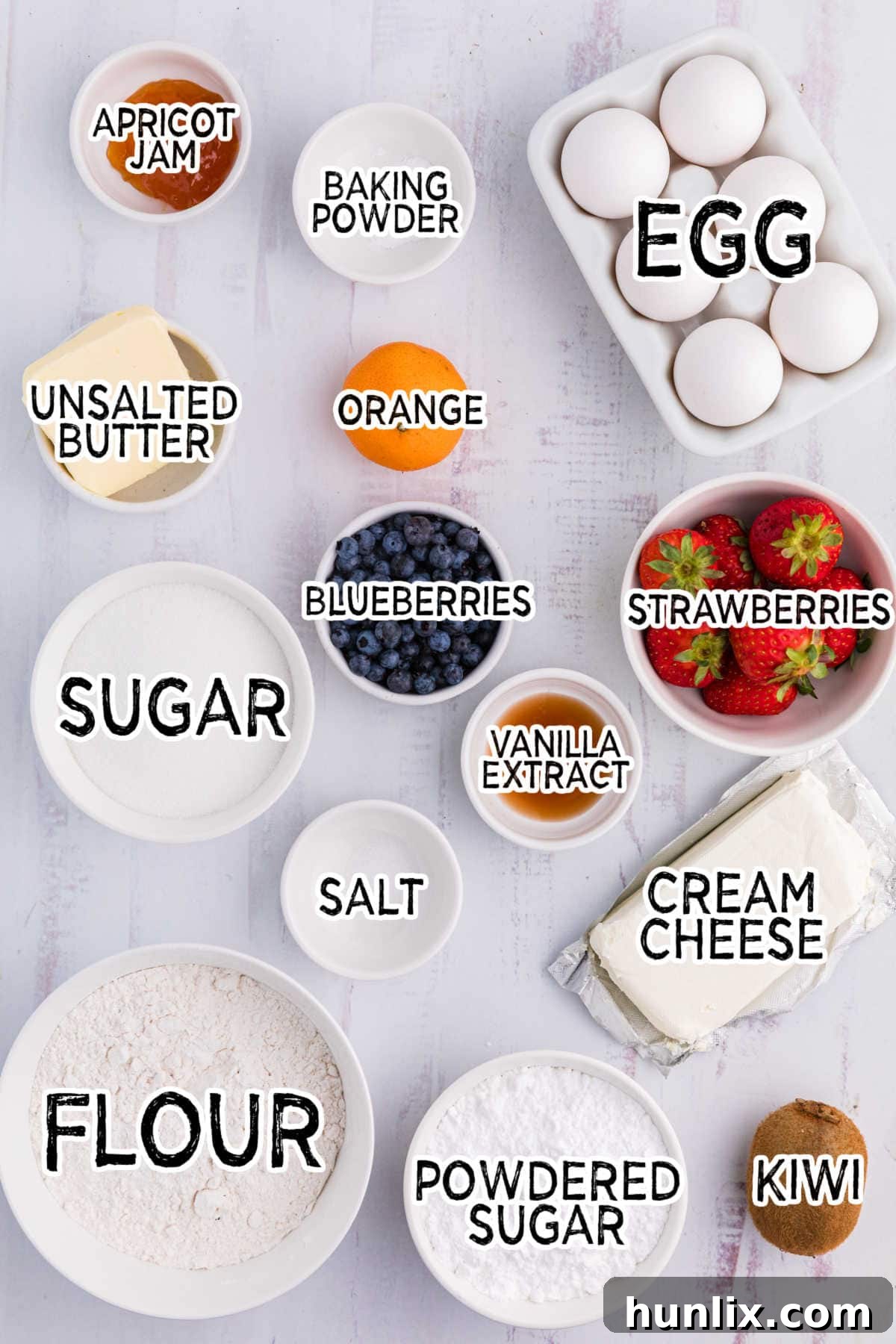 An assortment of fresh fruit pizza ingredients artfully arranged on a white background, including softened butter, granulated sugar, an egg, vanilla extract, flour, baking powder, salt, cream cheese, powdered sugar, sliced strawberries, whole blueberries, sliced kiwi, mandarin orange segments, and apricot jam, all ready for preparation.