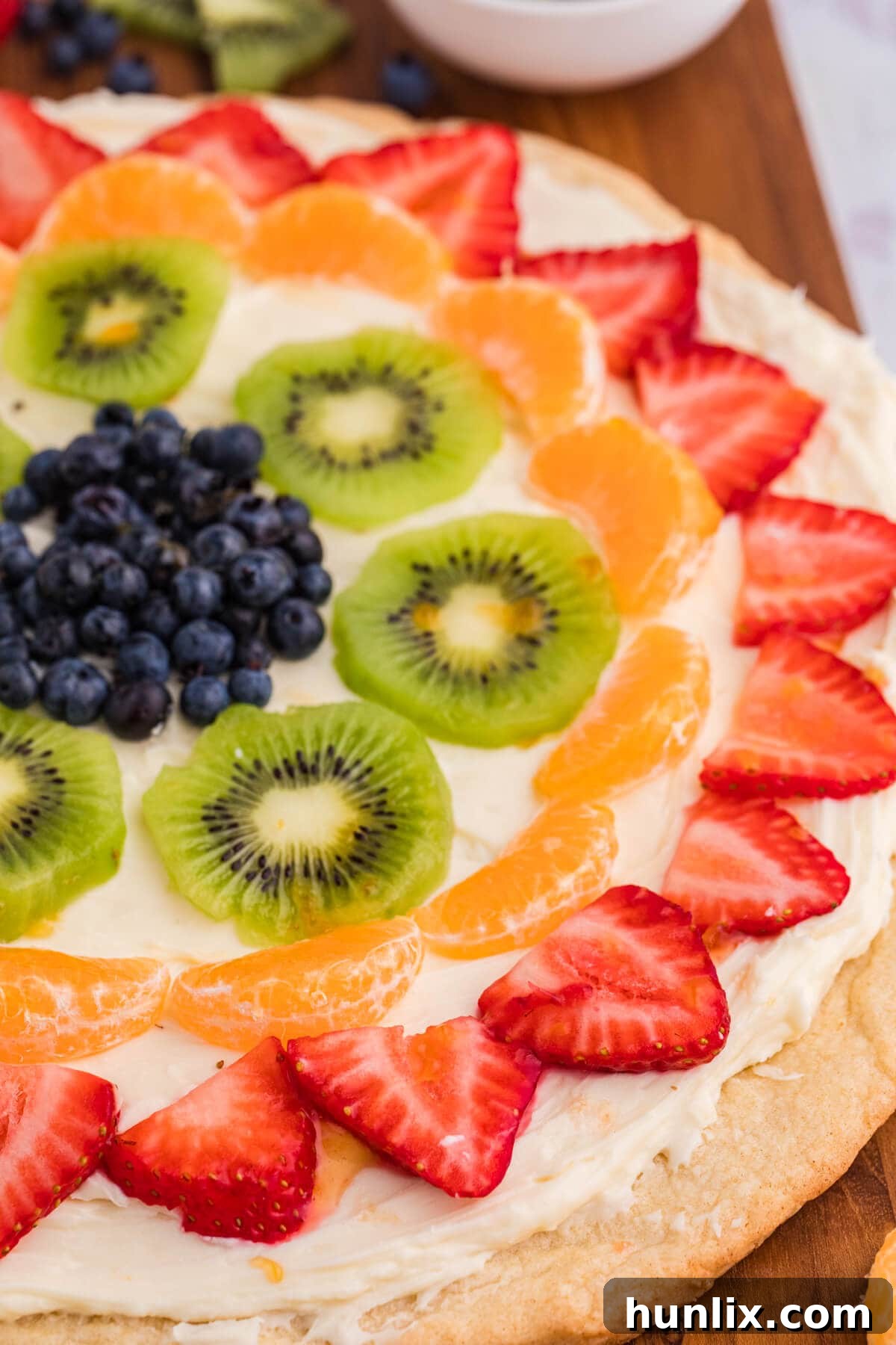 A close-up shot of a completed fruit pizza, showcasing its beautifully arranged fresh fruit, creamy frosting, and golden cookie crust, presented on a wooden board ready for slicing.