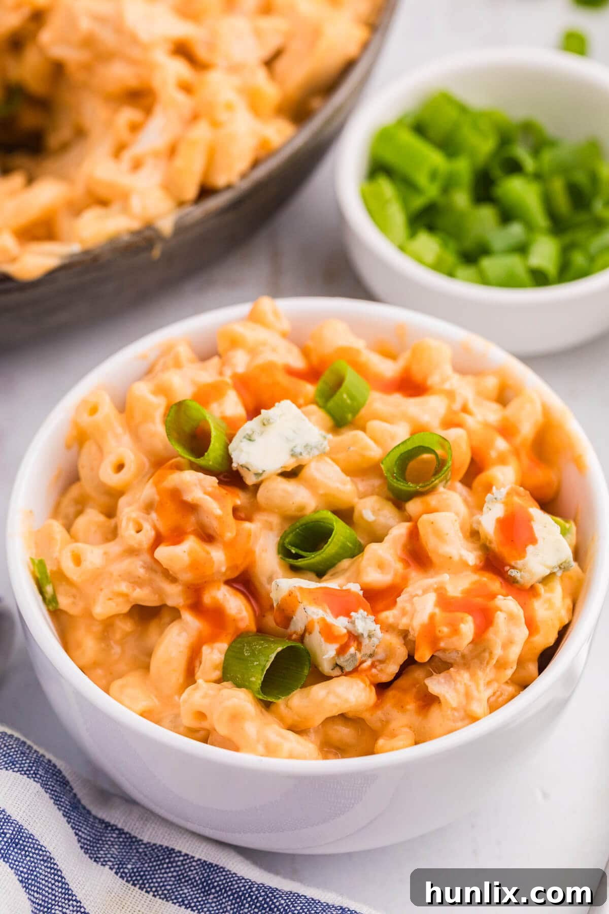 A generous serving of Buffalo Chicken Mac and Cheese, garnished with fresh green onions, presented in a clean white bowl on a textured background.
