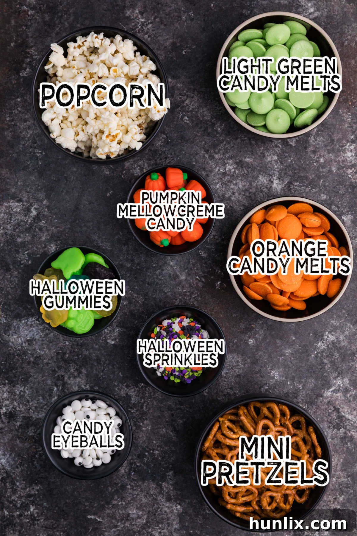 Spooky Halloween Popcorn 3 A close-up shot of Halloween Popcorn ingredients laid out on a dark surface, including popped corn, mini pretzels, pumpkin mellowcreme candies, green and orange candy melts, and candy eyeballs.