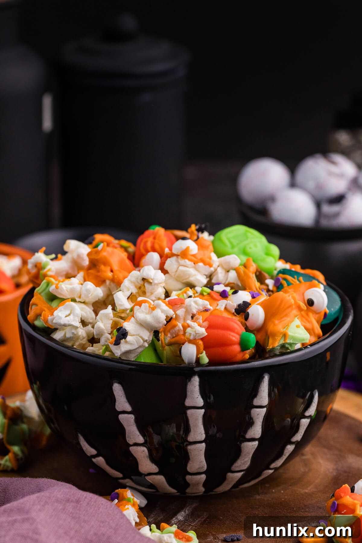 Spooky Halloween Popcorn 5 A bowl of Halloween Popcorn on a rustic wood board, showcasing orange and green drizzles, pretzels, mellowcreme pumpkins, candy eyeballs, and festive sprinkles.