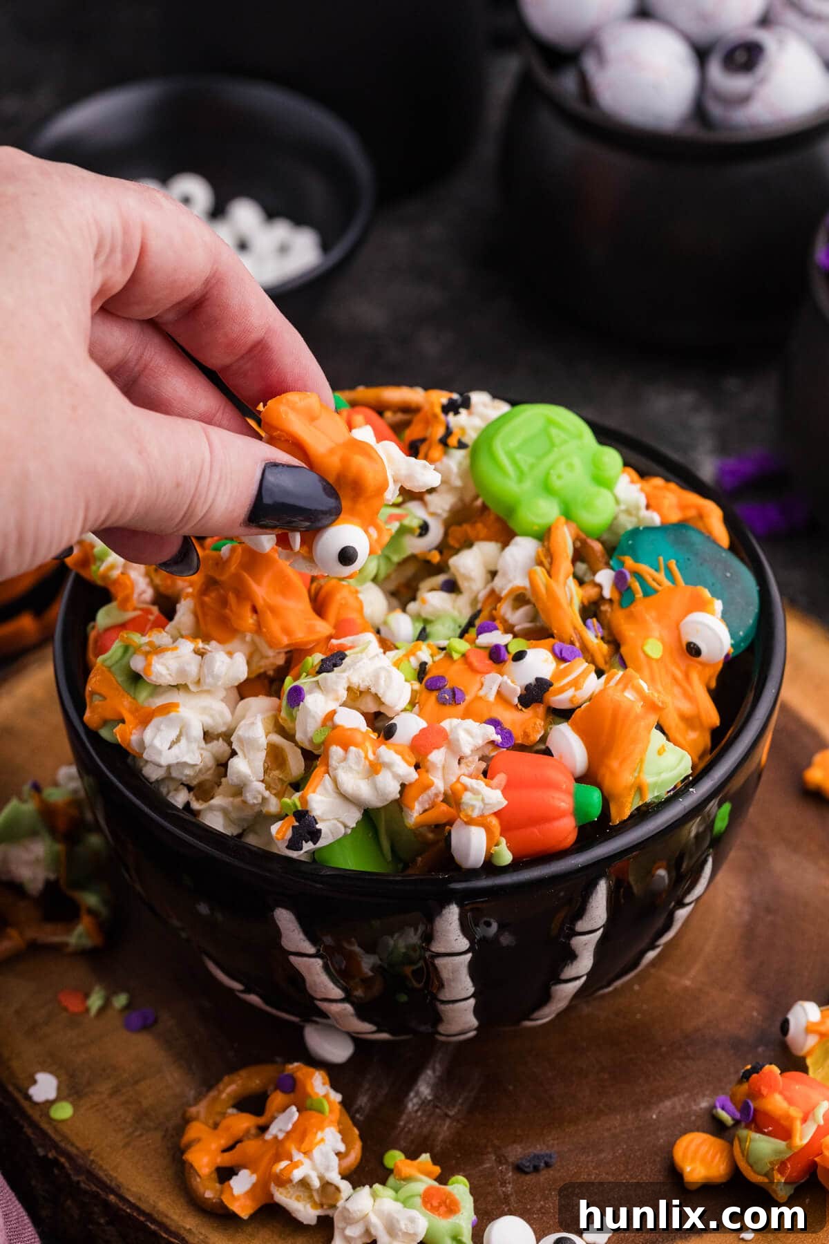 Spooky Halloween Popcorn 6 A hand lifting a cluster of Halloween Popcorn from a bowl, showcasing orange drizzle, popcorn, pretzels, a candy eyeball, and green gummies.