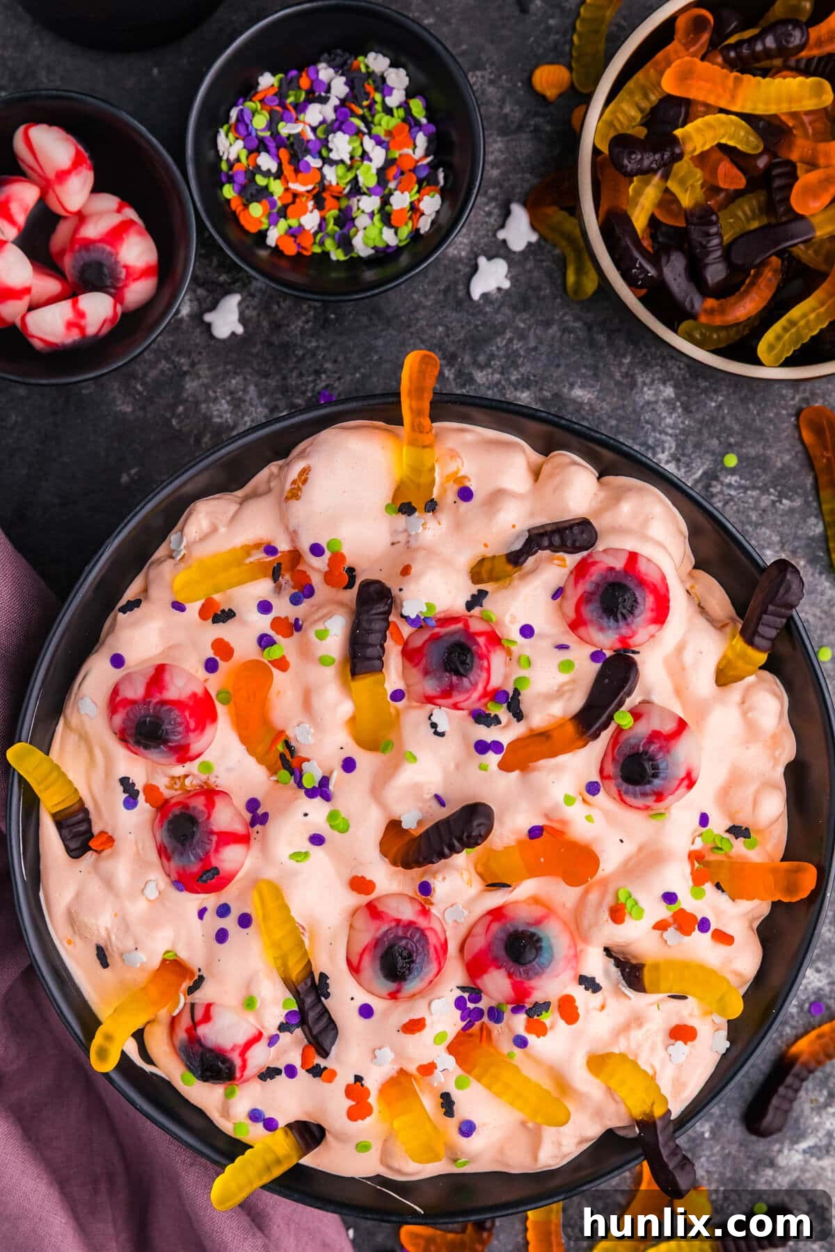 Overhead shot of a vibrant orange Halloween Fluff Salad, garnished with playful gummy worms, candy eyeballs, and festive Halloween sprinkles in a black bowl, set against a dark, spooky background.