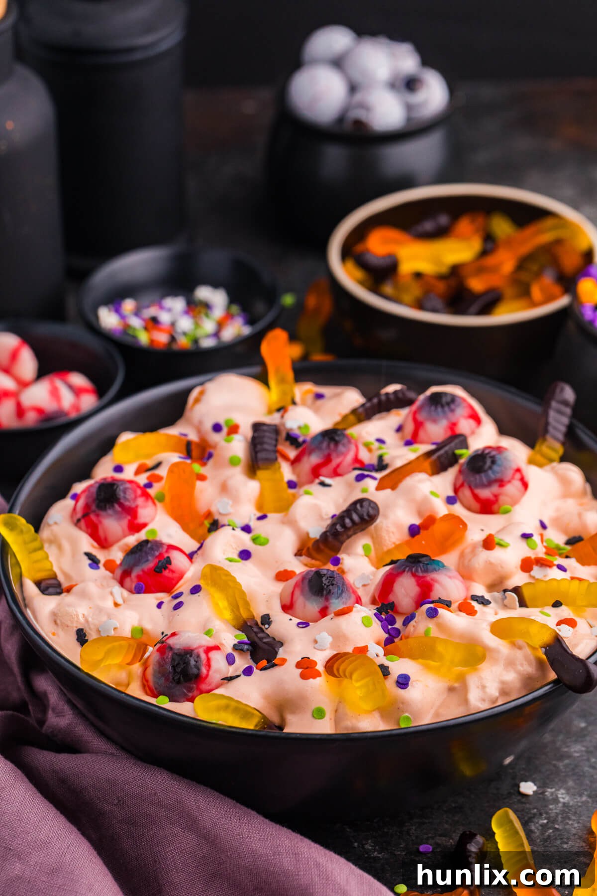 An angled view of a black bowl filled with vibrant orange Halloween Fluff Salad, artfully garnished with gummy worms and candy eyeballs. Smaller individual serving bowls are blurred in the background, suggesting a festive party setting or a buffet table.