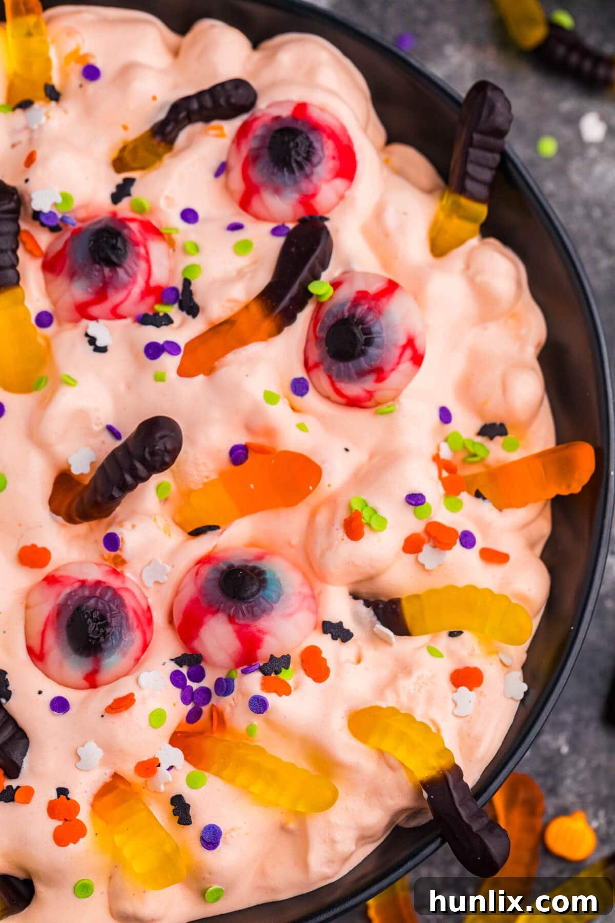 A vibrant close-up detail of orange Halloween Fluff Salad, meticulously topped with playful gummy worms, eerie candy eyeballs, and a medley of colorful Halloween sprinkles, highlighting its festive and inviting appeal.