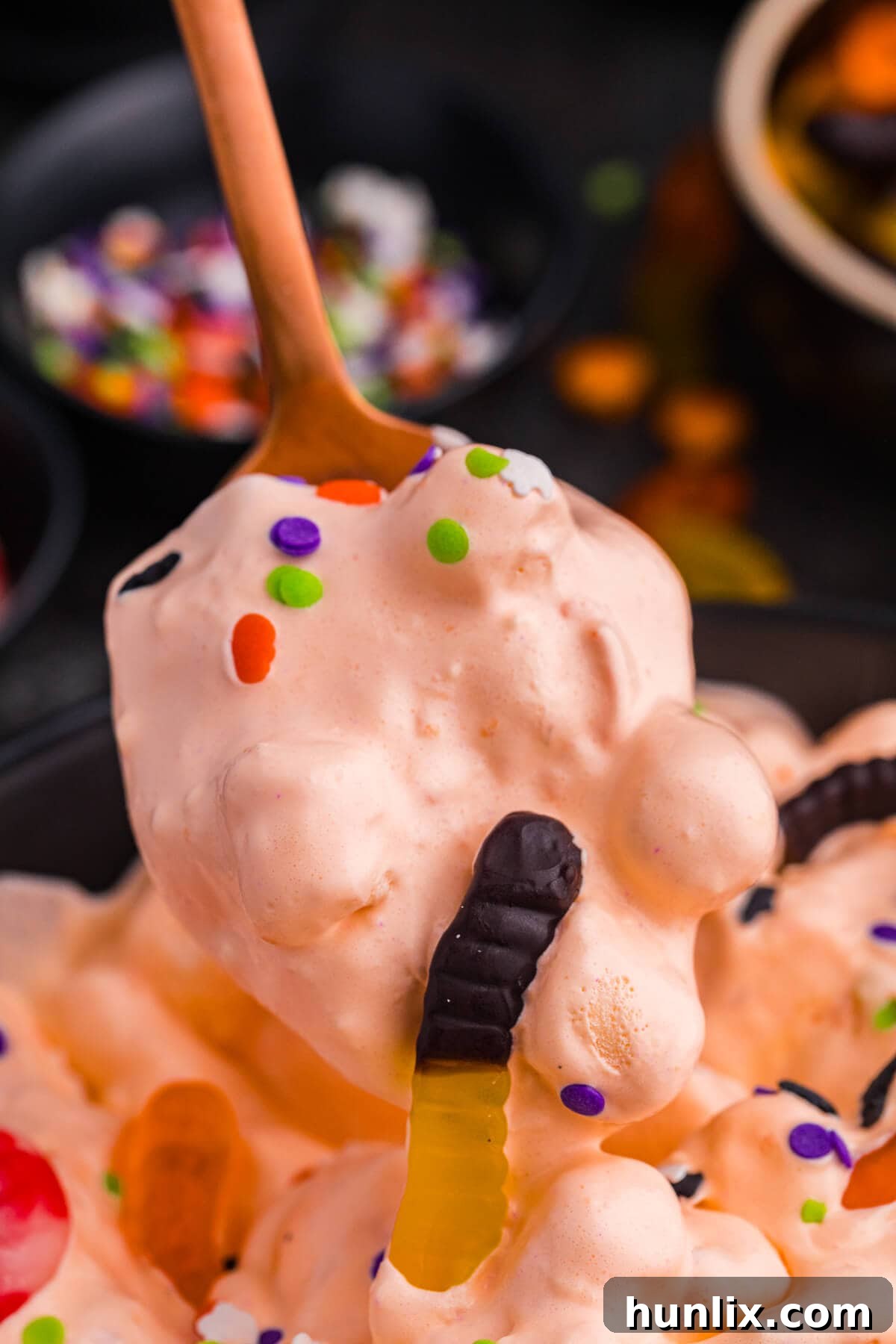 A spoon lifts a generous scoop of vibrant orange Halloween Fluff Salad, showcasing its airy texture, dotted with soft mini marshmallows, bright gummy worms, and festive Halloween sprinkles, ready to be enjoyed.