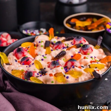 Wide hero shot of a black bowl brimming with Halloween Fluff Salad on a dark surface, surrounded by scattered gummy worms and small topping bowls in the background, creating a festive, spooky and inviting scene.